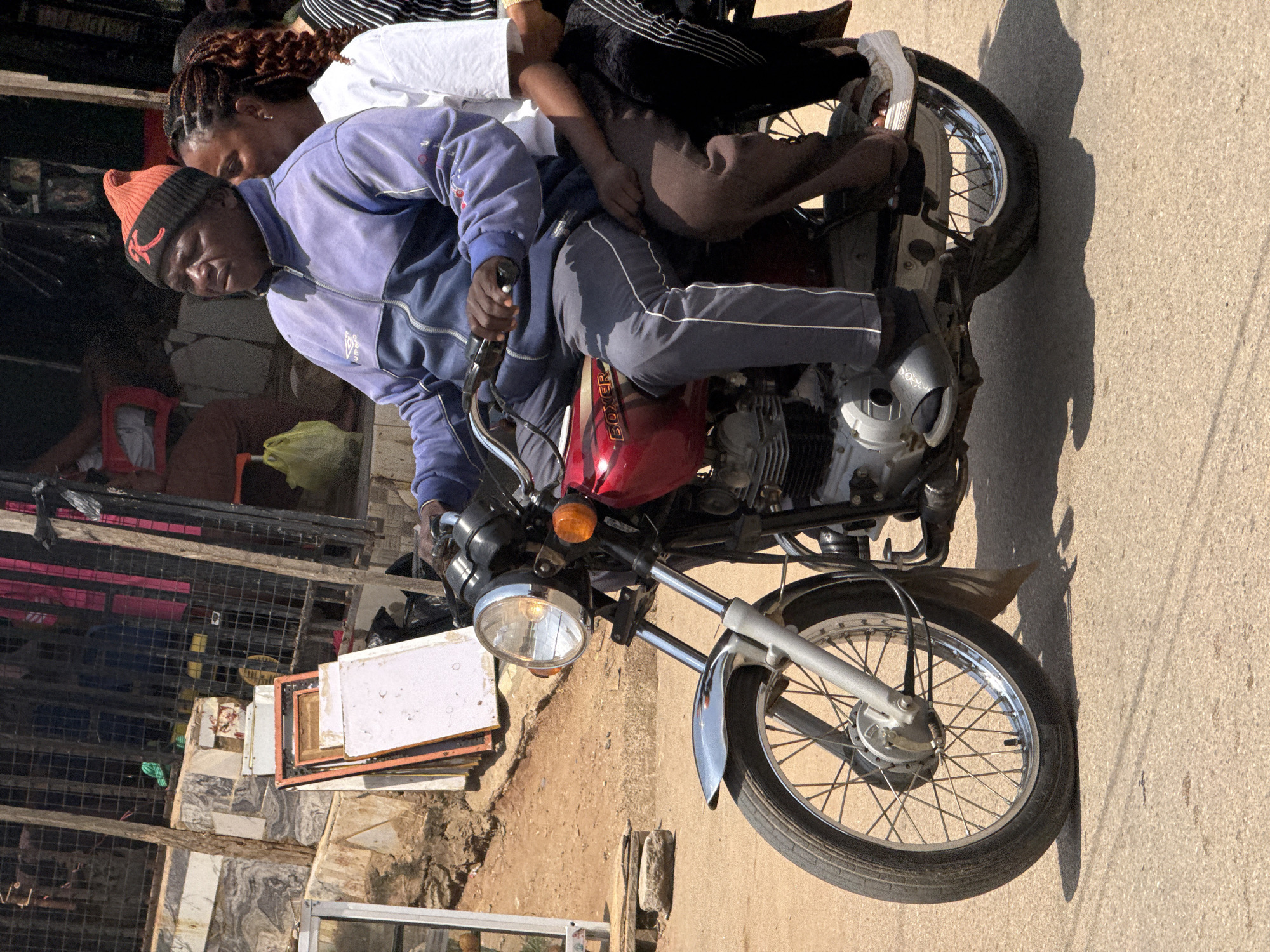 In Abuja, Nigeria, a man rides a red motorcycle with a woman passenger behind him. The man wears a blue jacket, grey pants, and an orange beanie with a black emblem. The woman is dressed in a white shirt and dark pants. They are on a paved road, with a market stall visible in the background. The stall has metal grates, stacked framed items, and various goods. The scene suggests a typical day in a busy urban area of Abuja.