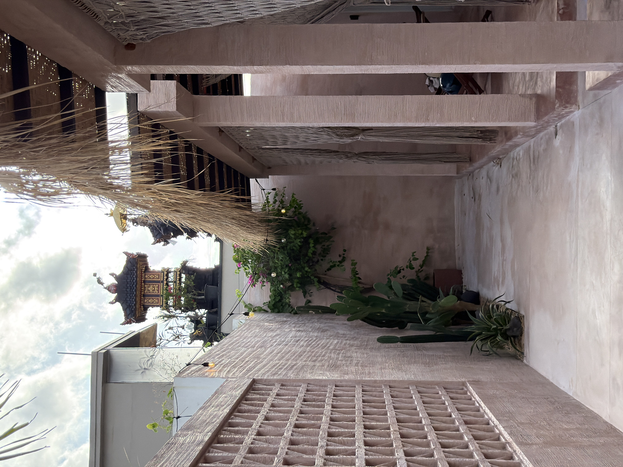 The image presents an upward-looking perspective within a building in North Kuta, Indonesia, likely a cafe or restaurant. The architecture features textured, light pink/beige walls and supporting structures constructed of solid rectangular blocks with hollow square vents. Various plants, including cacti and flowering vines, grow against the walls. Overhead, there is a woven, natural fiber roof structure. In the background, there is a glimpse of a building facade with a traditional Balinese shrine-like structure visible against a cloudy sky.