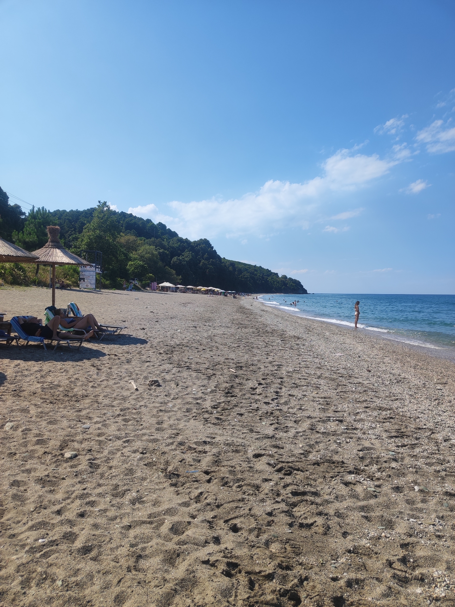 The image depicts a wide, pebbly and sandy beach in the Municipal Unit of Melivia, Greece, under a clear blue sky with scattered white clouds. The shoreline is lined with lush green trees and vegetation covering a hillside that stretches along the coast. The sea is a calm, bright blue. In the foreground, two people are relaxing on lounge chairs under a thatched umbrella. Further down the beach, several other umbrellas and lounge chairs are set up, with more people visible in the water and along the shoreline. A lone individual stands at the water's edge in the mid-ground. The overall scene conveys a tranquil beach atmosphere during daytime.