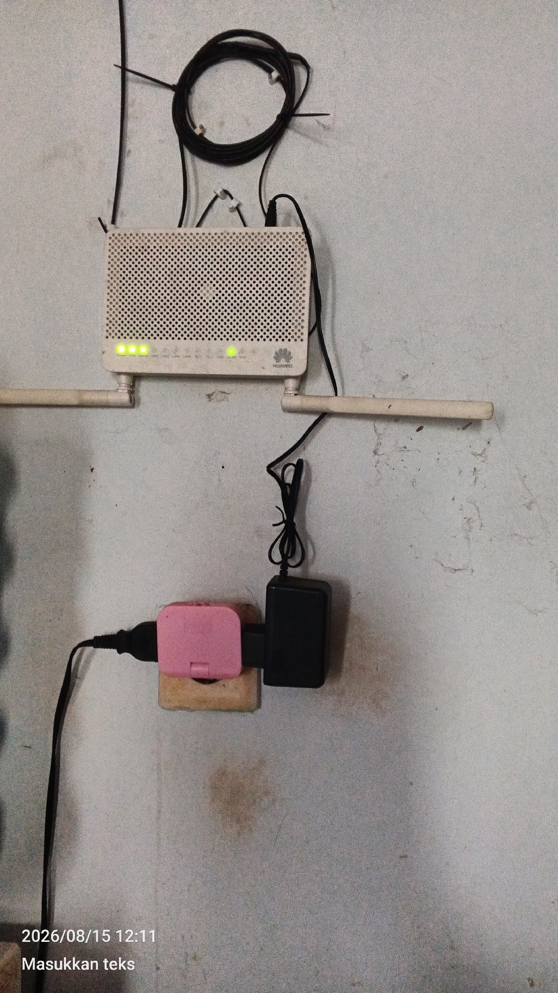 The image shows a white Huawei router mounted on a light-colored wall. Two white antennas extend from the router. A black power cord is connected to the router and runs down to a black power adapter plugged into a pink electrical outlet. The power adapter is then connected to a socket. There is a coiled black cable above the router, secured to the wall with white clips. The wall appears textured and dirty. In the bottom left corner, a timestamp reads "2026/08/15 12:11" followed by the text "Masukkan teks". The scene is indoors, likely in a residential or office setting in Jeru, Indonesia, given the Indonesian text.