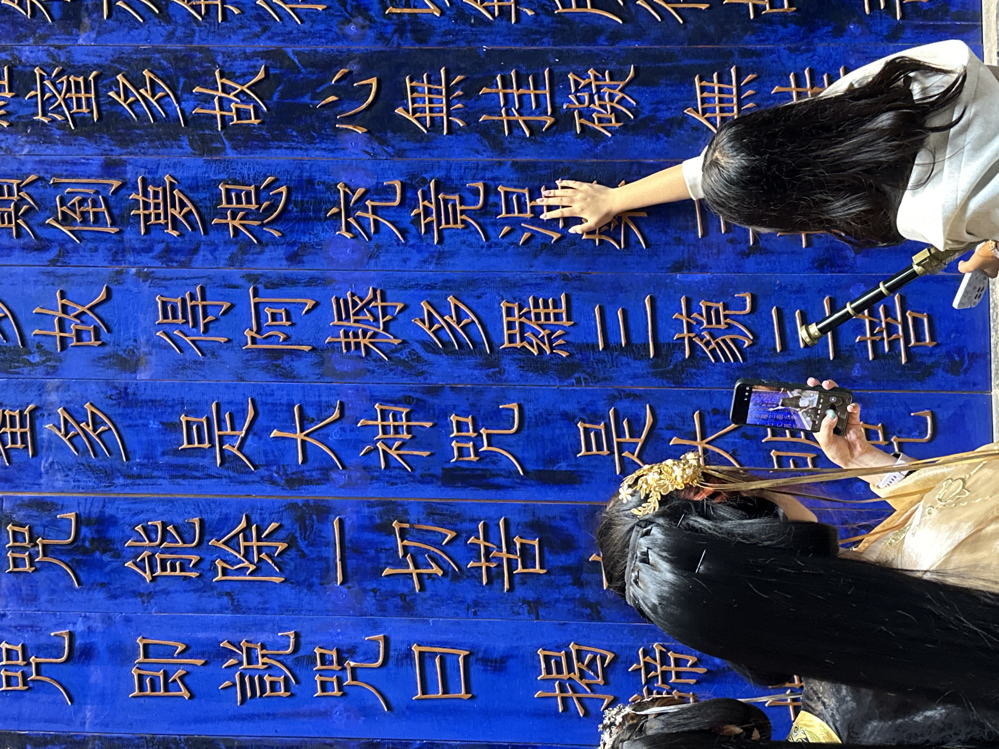 Here's a detailed description of the image:
**Scene:** The image depicts a scene featuring a large, blue wall with raised, gold-colored Chinese characters. Two people are interacting with the wall.
**People:**
* One person is touching the wall with their hand. They have long, dark hair and are wearing a light-colored top.
* Another person is taking a photo of the wall with their smartphone. This person has long, dark hair adorned with a golden decorative piece. They are wearing a golden-colored garment. A second person is below them wearing dark attire.
**Objects:**
* Large wall with Chinese characters.
* Smartphone.
* Decorative hairpiece.
**Actions/Interactions:**
* One person is touching the wall.
* The other person is using their smartphone to take a picture of the wall and the person touching it.
**Environment:** The blue wall suggests an indoor location. The scene is lit well enough to capture the details of the wall and people.
**Location (Implied):** The OCR provides a transcription of the Chinese characters. Given that the prompt asks for a location in Yuzhong District, China, it is probable that the scene depicts a cultural site or tourist attraction.