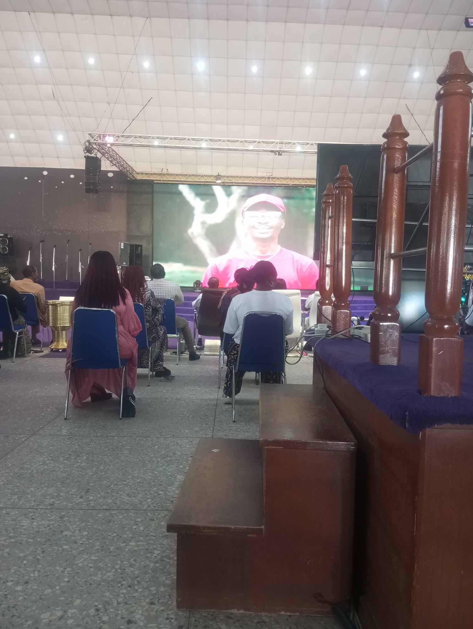 In Gura Top, Nigeria, this image captures a scene inside a large, well-lit indoor venue, likely a church or event hall, as indicated by the presence of a large screen displaying a person in a pink shirt and red cap, multiple rows of seating, and ornate wooden pillars. The floor is a speckled, light-colored terrazzo. Several individuals are seated, facing the screen. In the foreground, a person in a pink dress is seated on a blue chair, with another person in a white shirt to their right, also seated. In the mid-ground, more individuals are seated, with some in patterned clothing. To the left, microphones stand on poles, and a golden cylindrical object is visible. The overall atmosphere appears to be one of an organized gathering, with a focus on the presentation on the screen. The lighting is bright and even, suggesting artificial illumination.