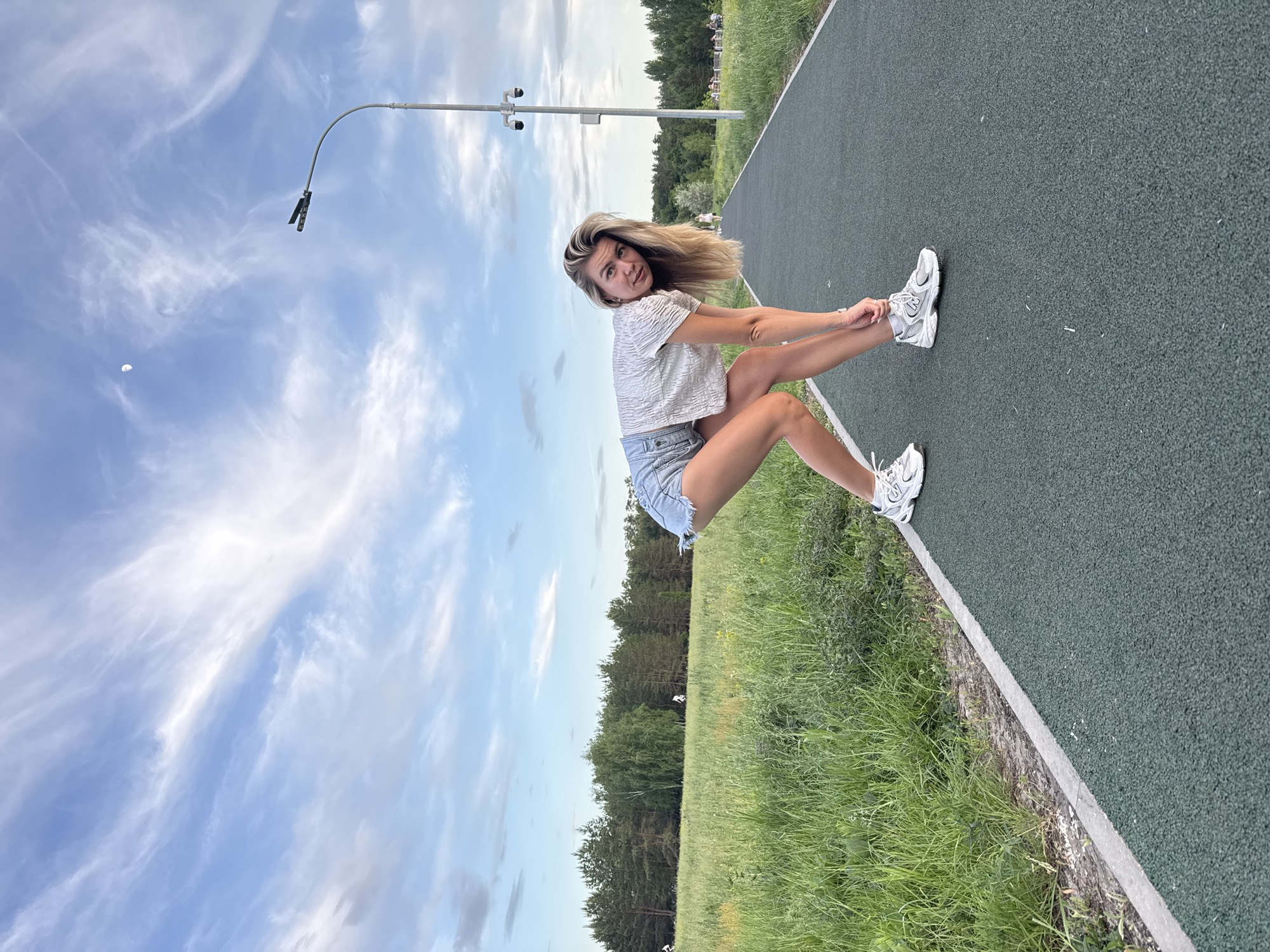 A woman with blonde hair, wearing a light-colored short-sleeved shirt and denim shorts, is bending over and tying the laces of her white sneakers on a green running track. The scene is outdoors, alongside a grassy area with trees in the background. A lamppost is visible in the background, and the sky is blue with wispy clouds. The location appears to be a park or recreational area.