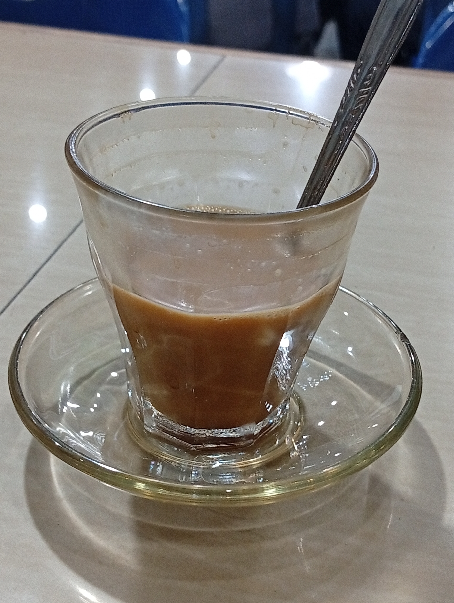 The image presents a close-up view of a partially consumed beverage in a clear glass cup, resting on a matching clear glass saucer. The cup contains a light brown liquid, indicative of coffee or tea mixed with milk or creamer, with some residue visible on the inner glass walls above the liquid line. A silver spoon, featuring an ornate pattern on its handle, is submerged in the drink.
The glassware is positioned on a light-colored, reflective surface, likely a table, which shows multiple bright, circular reflections from overhead light sources. Faint lines on the table suggest it might be a tiled surface. The background is softly blurred, revealing indistinct shapes and colors, including hints of blue and darker tones, possibly furniture or walls in an indoor setting.
The scene suggests a moment of pause or completion during a drink, likely in a cafe or an informal eatery. Given the provided context of Lhokseumawe, Indonesia, this setup is typical of a local coffee shop or warung serving popular beverages like *kopi susu*. No individuals are visible, and there is no discernible text or specific time of day indicated, beyond the artificial indoor lighting.
