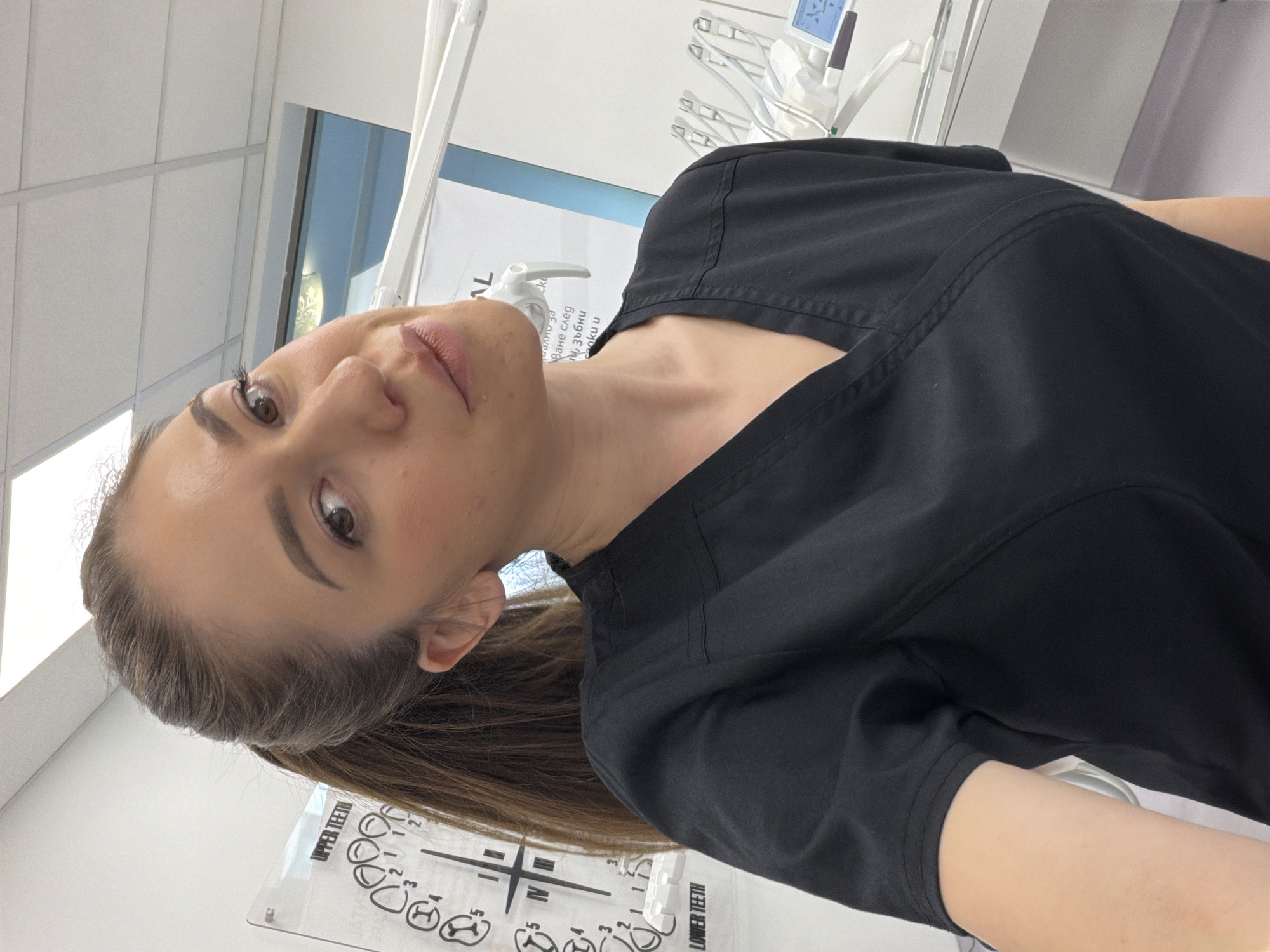 This is a portrait-style, slightly tilted, medium shot of a woman in a dental clinic. She is wearing black scrubs and her hair is pulled back. The woman is looking directly at the camera with a neutral expression. In the background, there are dental instruments and a chart with diagrams of teeth labeled "UPPER TEETH" and "LOWER TEETH". The overall setting is bright and appears to be a professional medical environment. The text on the chart and a nearby sign indicate the context is related to dentistry. The sign also contains some Cyrillic text.