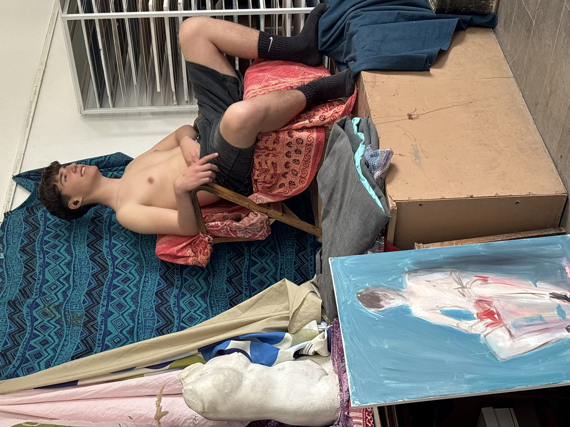 A person is lying on a blue patterned textile on the floor with their head near a white wall and a metallic framed window. The person is wearing dark shorts and black socks with a white logo. The legs are propped up on a small, red patterned chair or stool. A brown cardboard box and a blue-painted canvas with a figure sketched on it are located near the chair. Other objects, including what appears to be a white statue or bust, are scattered around the floor. The location is not specified.