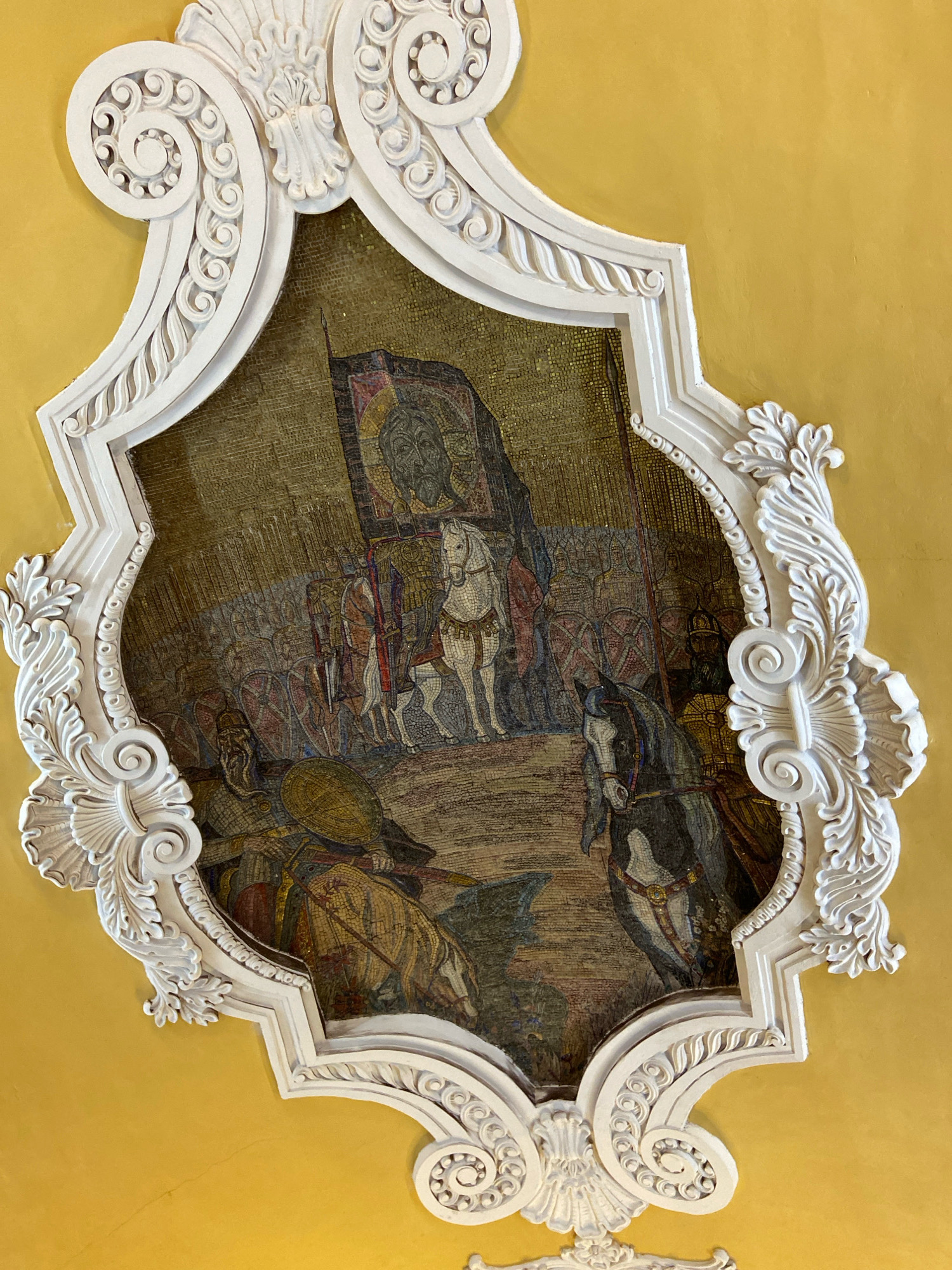 This is a detailed description of a mosaic artwork set within an ornate white stucco frame on a yellow ceiling, likely located in the Moscow Metro. The mosaic depicts a historical battle scene. In the foreground, a fallen warrior in armor, wearing a helmet and holding a large round shield, lies on the ground. To the right, a dark-colored horse with a rider in armor is depicted. Behind them, a procession of soldiers with shields and spears is visible, with a large banner held high at the center. The banner features a dark, stern face, possibly religious in nature. The background is rendered in a mosaic of golden and brown hues, creating a sense of depth and historical texture. The white stucco frame is highly decorative, featuring scrollwork, acanthus leaves, and geometric patterns, characteristic of Neoclassical or Baroque architectural styles often found in Moscow's metro stations. The overall scene evokes a sense of ancient warfare and heroic struggle.