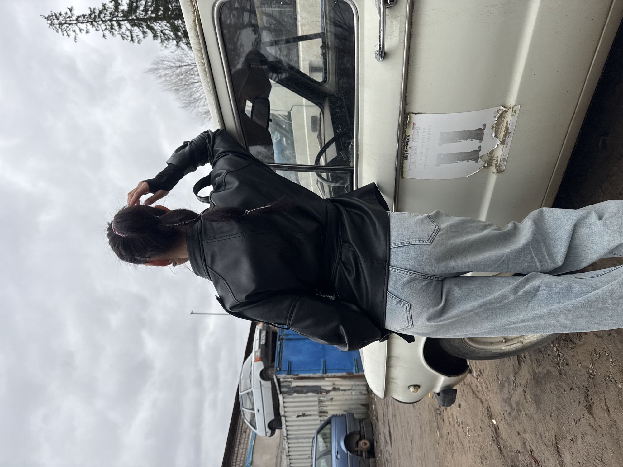 A vertical eye-level shot shows a person in a black leather jacket and blue jeans standing next to an old, white car. The person is facing away from the camera, with their right hand touching their hair. The car has a worn sticker on its side with illegible writing and partially obscured images. The car's window is partially open, revealing the interior with a steering wheel. In the background, there are other cars, a blue structure resembling a garage or shed, and a cloudy sky with bare trees. The ground appears to be a dirt or gravel surface. Based on the location in Chaikovsky, Russia, as indicated in the image parameters, the scene potentially represents a parking area or junkyard with aging vehicles and industrial elements.