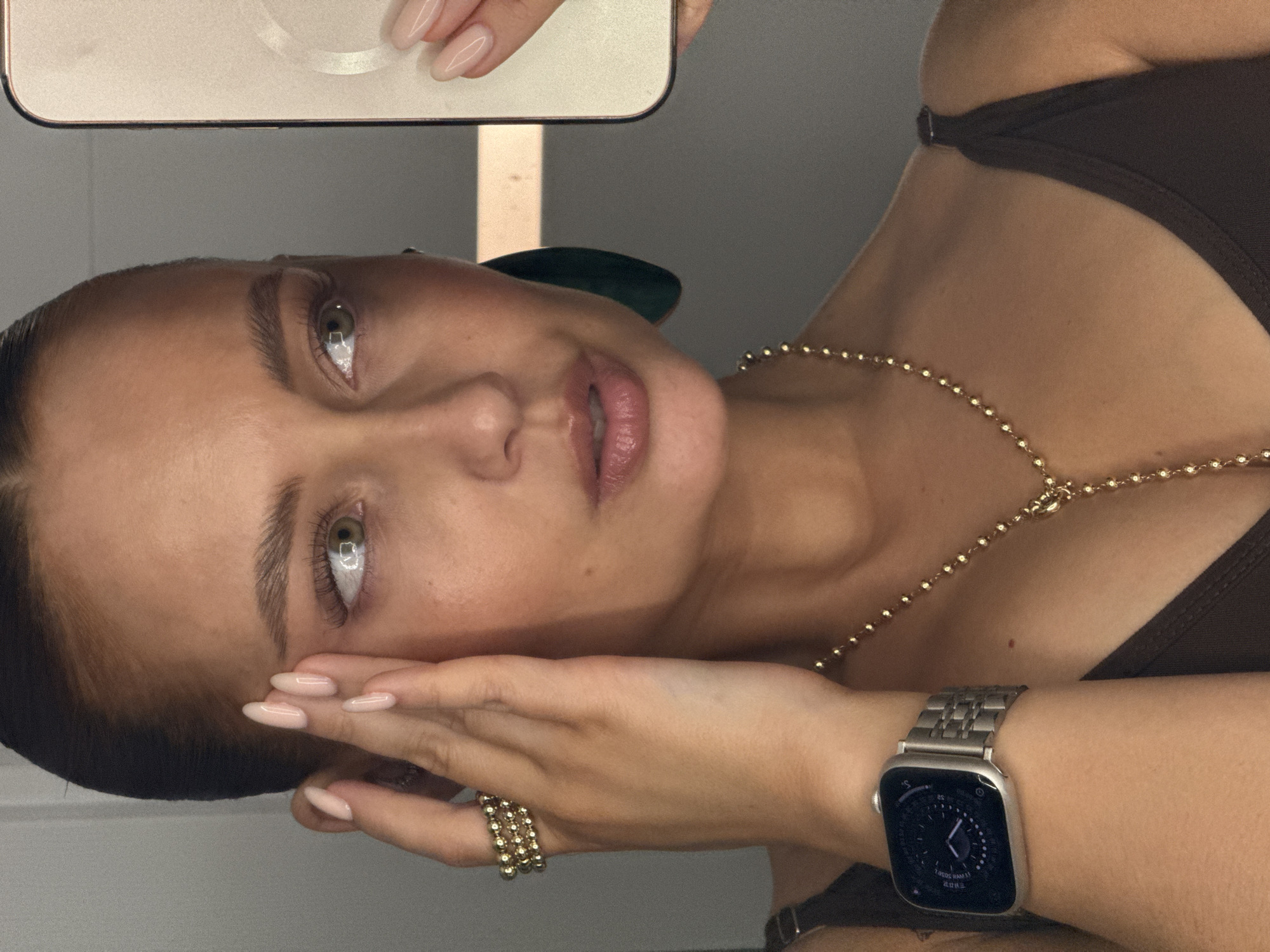 A woman is holding a smartphone in her left hand, taking a selfie. Her right hand is near her face, adorned with a gold ring and an Apple Watch with a silver band. She is wearing a brown top and a gold necklace. The background is a light gray wall. There is no indication of this picture being taken in Chaikovsky, Russia.