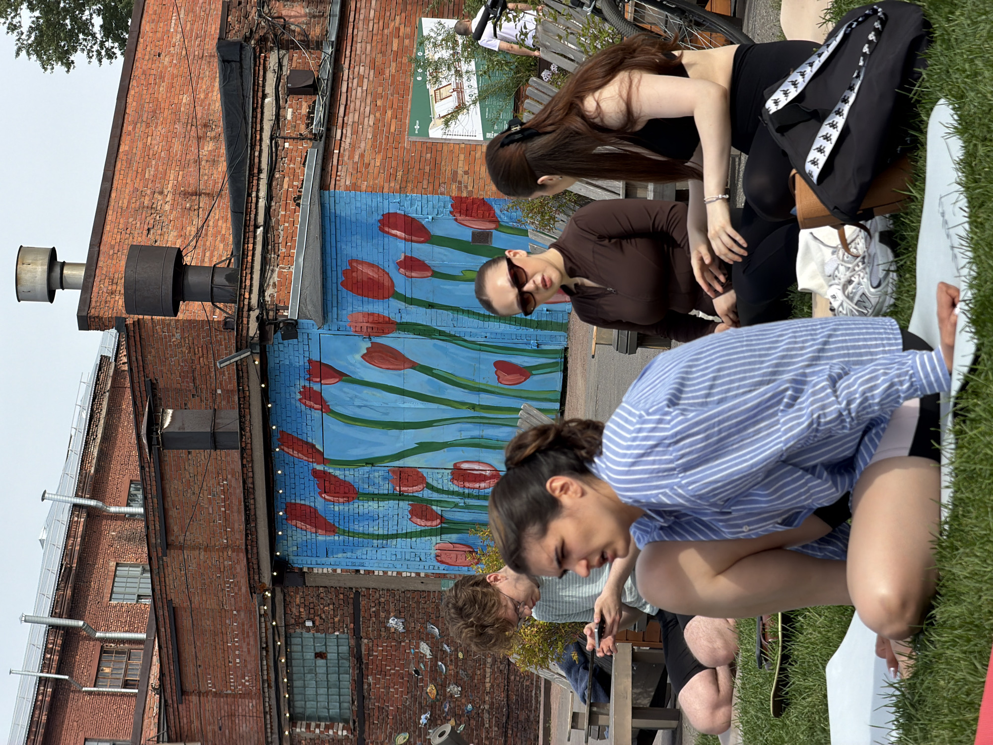 The scene depicts an outdoor leisure area within a repurposed industrial complex in Saint Petersburg, Russia. In the foreground, three individuals are seated on a grassy lawn.
On the left, a woman with dark hair in a bun is seated on a white mat, wearing a blue and white striped long-sleeved shirt and black shorts with a white band. She faces slightly left with her mouth open. Behind her, a young man with light brown, curly hair and glasses is seated on a wooden bench, engrossed in a smartphone he holds. He wears a light striped t-shirt and dark shorts, his legs bare on a white mat.
In the center-right foreground, two women are seated on beige mats. The woman on the left wears dark sunglasses, a dark brown zip-up long-sleeved top, and dark pants. She looks towards her right with her mouth slightly open. Facing her is another woman with long, dark brown hair styled half-up with a black clip, wearing a black bralette-style top and black leggings. Her hands are clasped in front of her. Near them, a black tote bag with a white and black "Kappa" logo strap, white and grey New Balance sneakers, and a small brown bag are placed on the grass.
The background features several red brick buildings. A prominent blue mural with large red tulips and green stems covers a section of a building wall or large door. Above this, string lights are visible. The buildings exhibit various architectural elements, including windows with grids, several metal exhaust pipes, and large cylindrical structures on the rooftops, one in silver and another in black. On the far right, a green information board with a diagram is mounted on a wall, and a man in a white t-shirt is partially visible walking behind a low wooden barrier. A bicycle is also partially visible on the far right. The sky overhead is light grey.