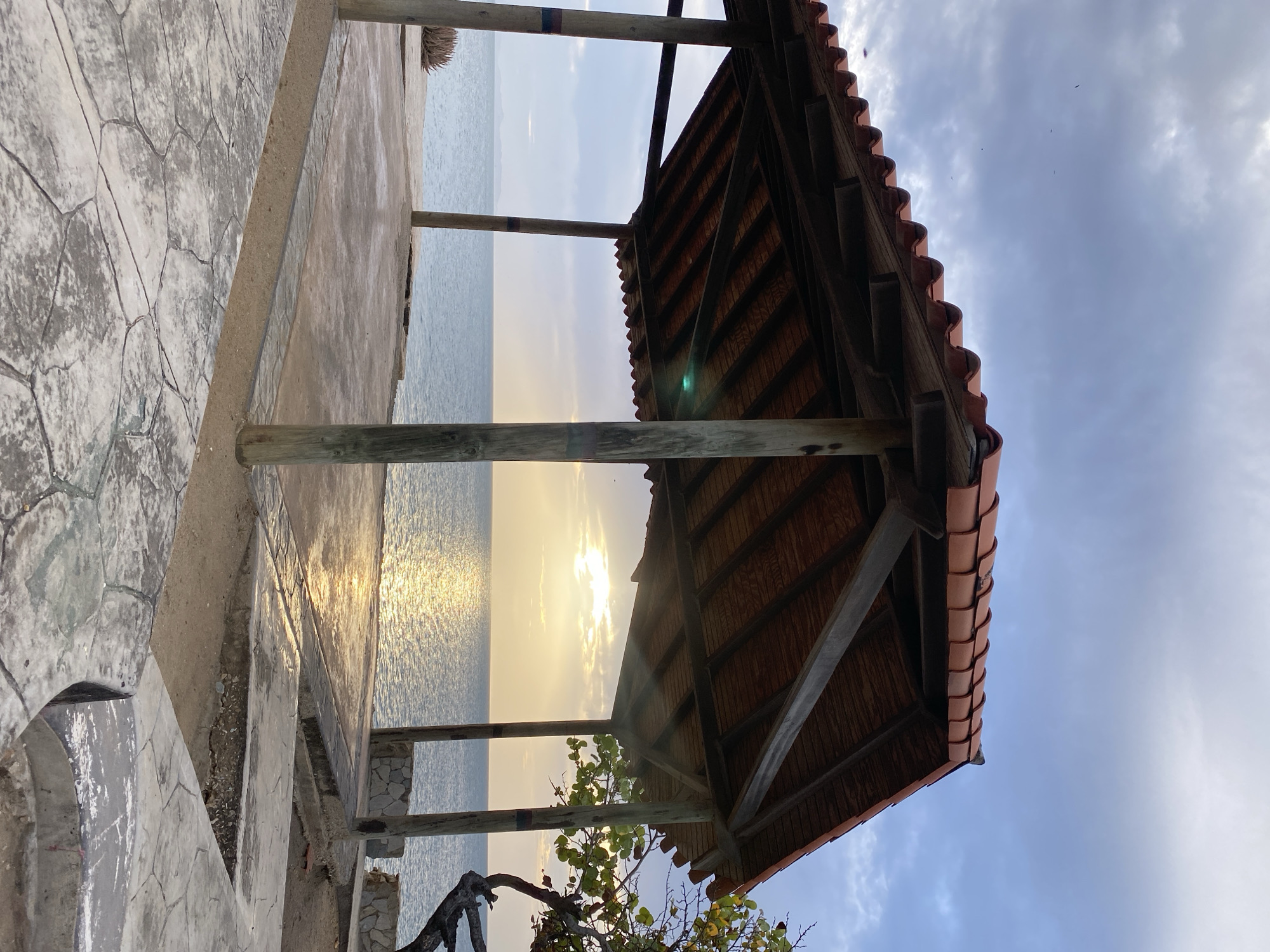 This image depicts an open-air, covered structure with a tiled roof and wooden supports, situated on a paved area overlooking a body of water. The structure appears to be located in Juan Griego, Venezuela, with the Caribbean Sea visible in the background. The sun is setting, casting a golden reflection on the water's surface. The surrounding environment includes a tree with dark branches on the right side of the frame and stone-paved pathways in the foreground. The sky is cloudy with some patches of blue. There are no people visible in the scene.