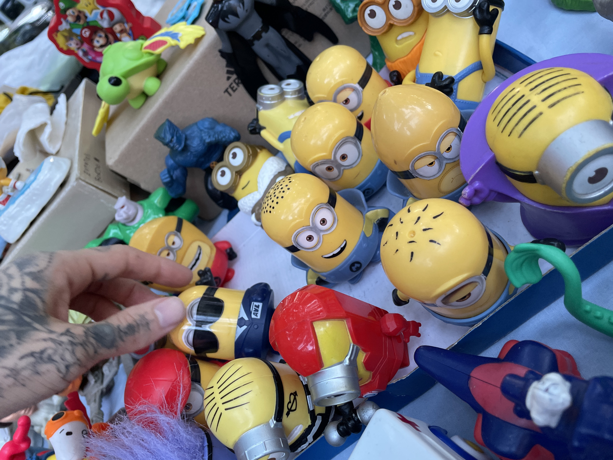 The image depicts a collection of toys, primarily Minions figurines, scattered in a cluttered manner. Some other toys are present, including a green turtle, a blue gorilla, and what appears to be a Batman figure. The objects are arranged in a haphazard way, suggesting a disorganized collection. A tattooed hand reaches into the pile of toys. The image has no clear indication that would denote it's located in Buenos Aires, Argentina.