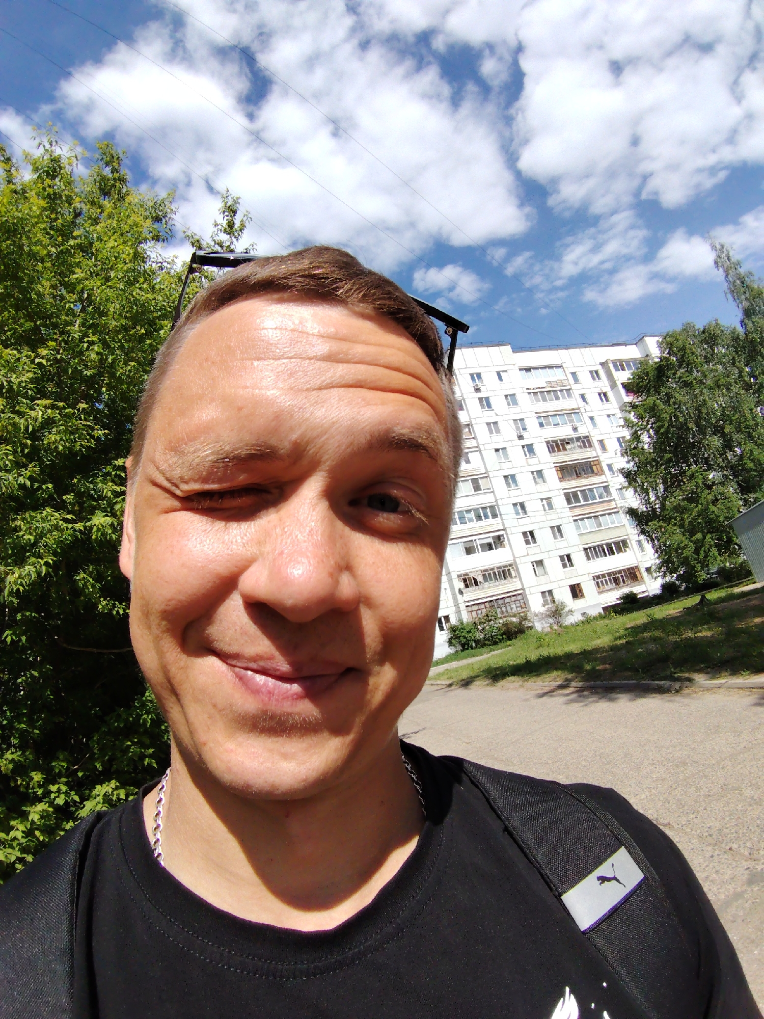 A close-up, selfie-style photograph depicts a smiling man with one eye closed in a wink, wearing a black t-shirt and a silver chain. Sunglasses are positioned on top of his head. The background reveals an urban environment, specifically the city of Chaikovsky, Russia. A multi-story apartment building, painted white with visible windows, stands behind him, partially obscured by trees with green foliage. The sky is visible above, displaying a mix of blue and scattered white clouds. A paved road or pathway is in the lower foreground. A Puma logo is visible on the backpack strap on the man's shoulder.