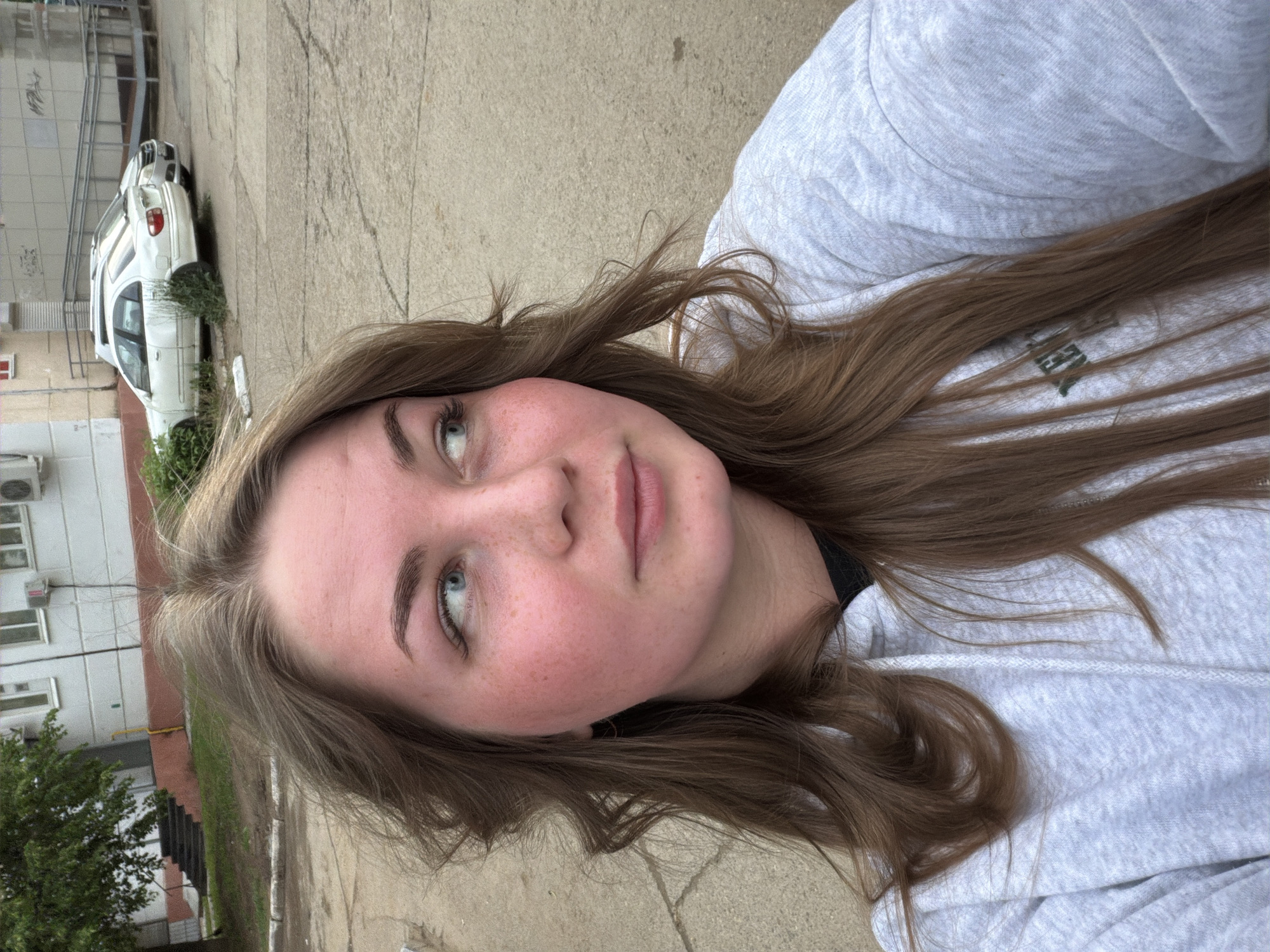 A young woman with long brown hair and blue eyes is depicted in an outdoor setting. Her fair complexion shows some redness, possibly sunburn or rosacea. She is wearing a light gray hooded sweatshirt. The background reveals elements of a city environment: a light-colored building with air conditioning units, a parked white car, and concrete pavement. The scene suggests a location in the city of Chaikovsky, Russia. The angle of the photograph indicates that it was likely taken as a selfie.