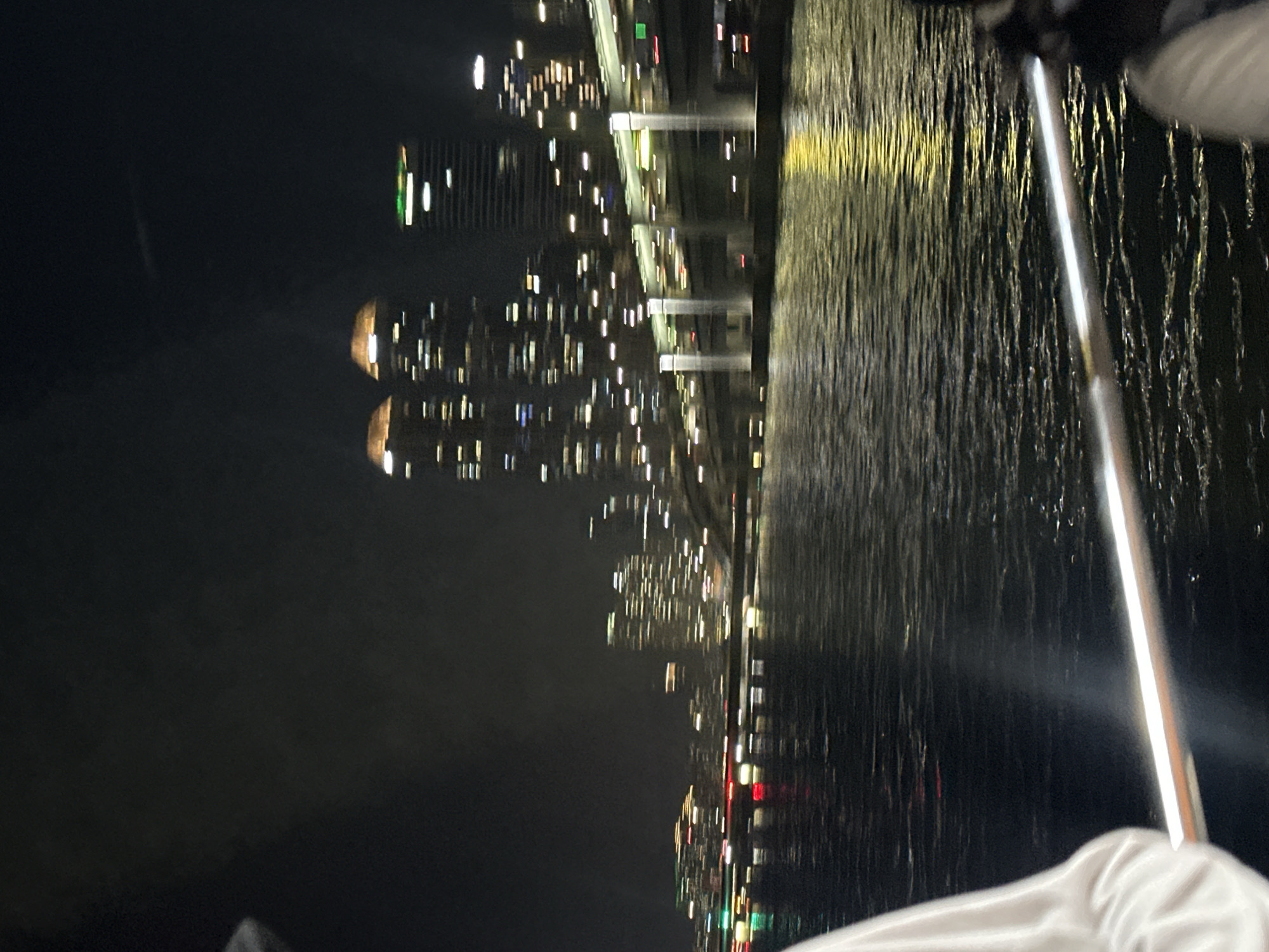 The image captures a nighttime view of the city of Busan, South Korea, likely from a boat on the water. In the background, a city skyline is visible with numerous illuminated buildings and skyscrapers. A large bridge with lights spans across the frame, with vehicles appearing as streaks of light, indicating movement. The water in the foreground reflects the city lights and the bridge lights, creating a shimmering effect. In the lower left corner, the shoulder and arm of a person wearing a white garment are visible, and in the lower right, a portion of another person's head and shoulder are visible. The overall scene conveys a sense of urban nighttime activity along a waterfront.