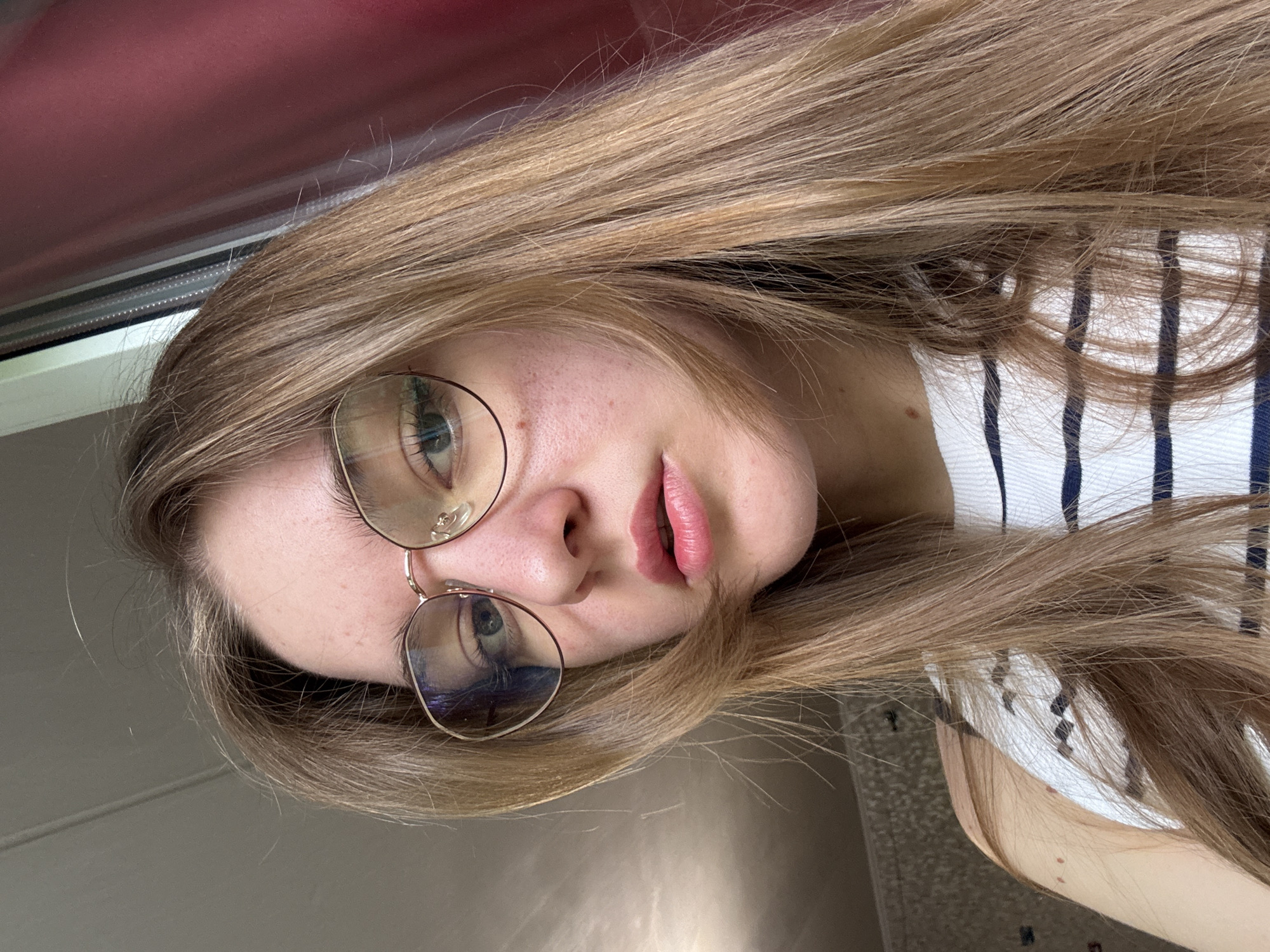 A woman with long, light brown hair is wearing glasses and a white and navy striped shirt. The photo appears to be taken indoors, possibly near a window with a red curtain. The city of Perm, Russia, cannot be confirmed or refuted from the image.