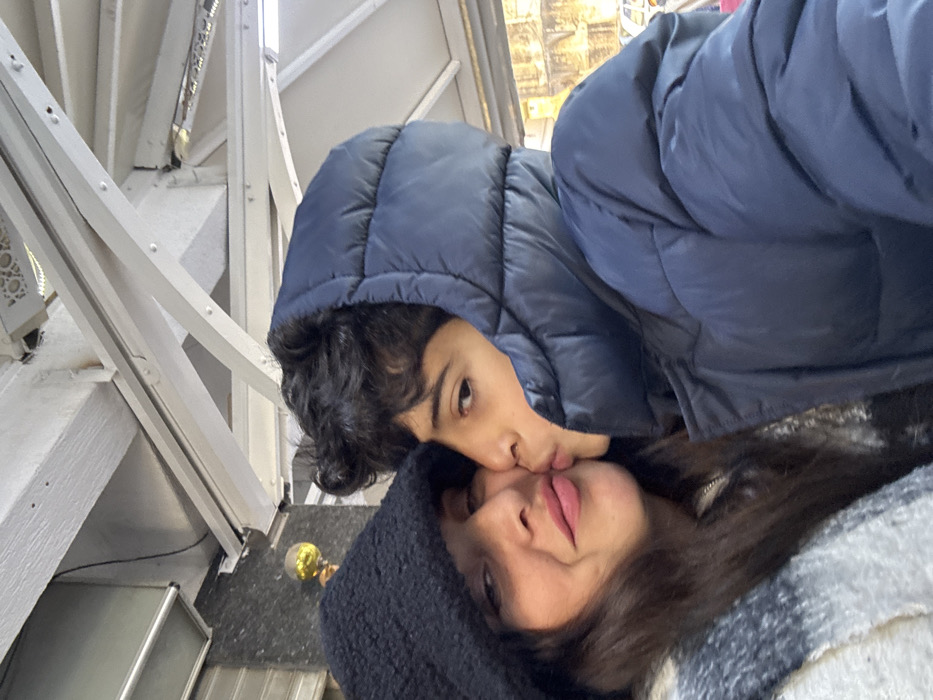 The image displays two individuals in a close-up, outdoor setting within the city of Azrou, Morocco. On the left, an adult female with dark hair is visible, wearing a black fuzzy hat and a grey, black, and white plaid scarf. Her lips are slightly puckered, adorned with light pink lipstick. To her right, a younger male, wearing a dark blue hooded puffer jacket with the hood up, is kissing the female on her left cheek. He has dark, curly hair and his eyes are open, directed forward. The background features a white overhead structural framework composed of beams and cross-members, with small rivets visible. To the upper left, a dark, possibly tiled or stone wall section is present, along with a golden spherical object and a partially visible metallic roll-down shutter. The upper right background shows a decorative white panel on the overhead structure, while the far right reveals blurred, lighter-toned architectural elements.