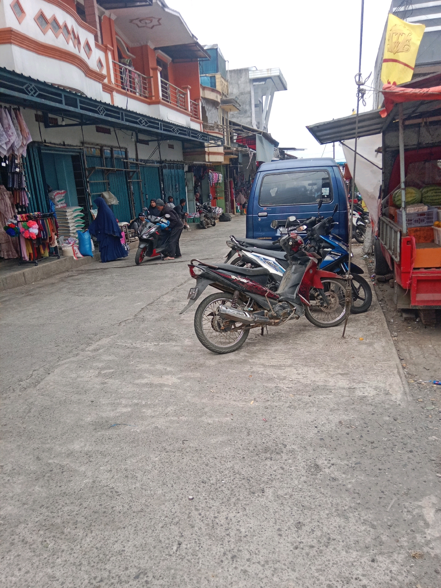 The image captures a street scene in Batur, Indonesia, likely a market area. In the foreground, a gray asphalt road is visible, showing signs of wear and tear. Several motorcycles are parked or are being interacted with. A red and black motorcycle is prominently displayed in the middle of the frame. To its left, a blue van is parked. Further back, a woman in a blue hijab is walking past a stall displaying clothing on racks. Several other individuals, some appearing to be vendors or customers, are seen near the stalls and motorcycles. The stalls themselves are covered by awnings and appear to be selling various goods, including textiles and possibly food items, with some sacks of what might be rice or other dry goods stacked near one of the stalls. In the background, buildings with multiple stories line the street, featuring orange and white facades and balconies. A red truck with a load of watermelons is parked to the right, adjacent to a stall. The sky is overcast, suggesting a cloudy day. A yellow flag with an emblem is visible on a pole on the right side of the image. The overall impression is that of a busy local marketplace.