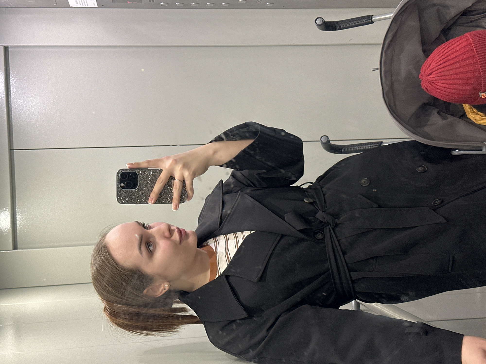A young woman is taking a selfie in a mirrored elevator, likely in Chaikovsky, Russia, though this cannot be definitively confirmed visually. She is wearing a black trench coat and holding a cell phone with a glittery case. A baby stroller with a red knit cap inside is partially visible to the right of her. The elevator interior is plain with light gray walls.