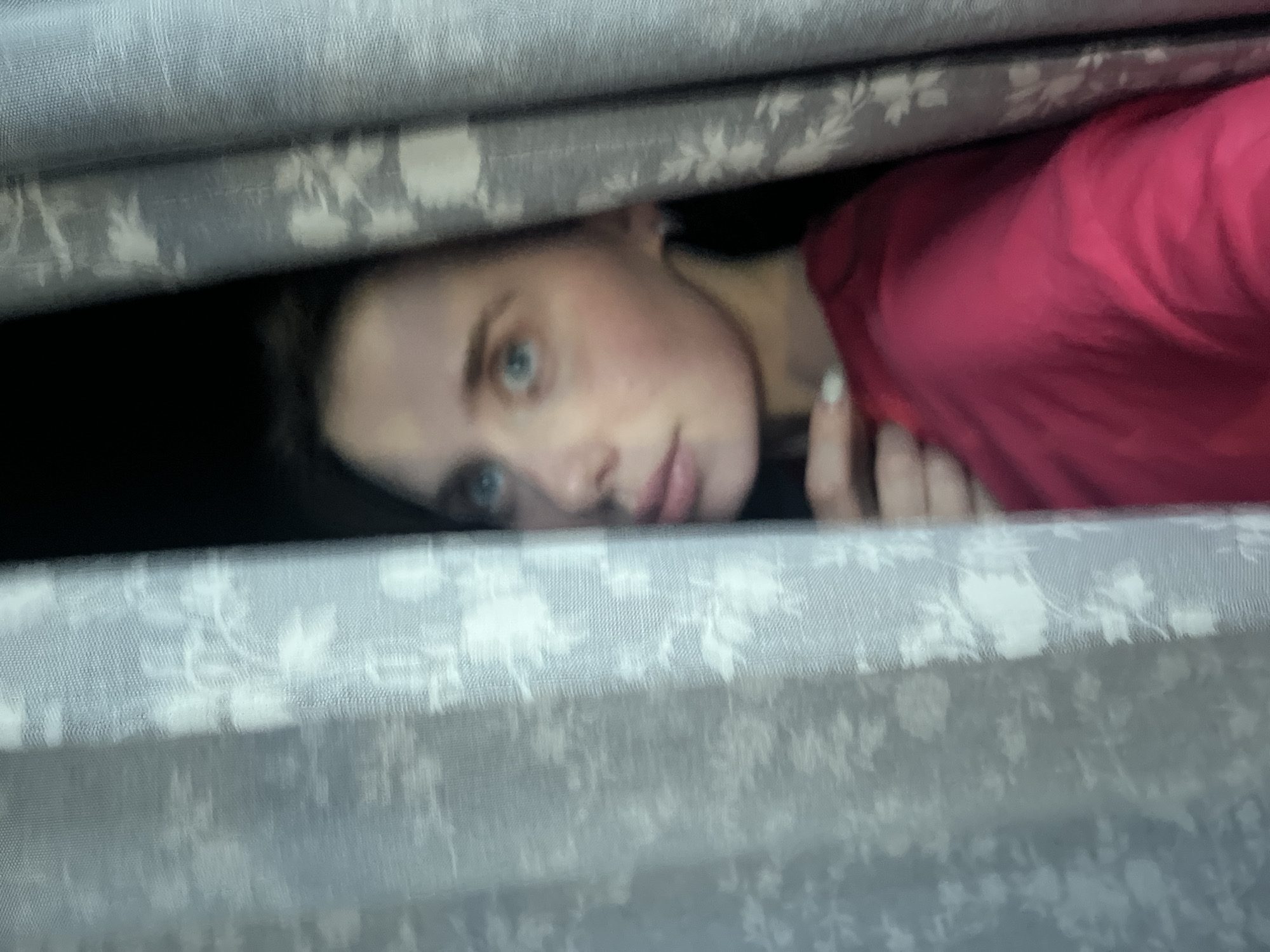 A young individual is partially visible, peeking from behind two light grey curtains adorned with a repeating white floral pattern. The subject, displaying light skin, blue eyes, and dark hair, is positioned centrally, looking upwards and to the viewer's left. A hand with light-colored nail polish is visible, holding the right curtain open. The individual wears a bright red or deep pink garment. The background behind the subject is dark, indicating an unlit space or exterior. This scene is observed within the city of Chaikovsky, Russia.