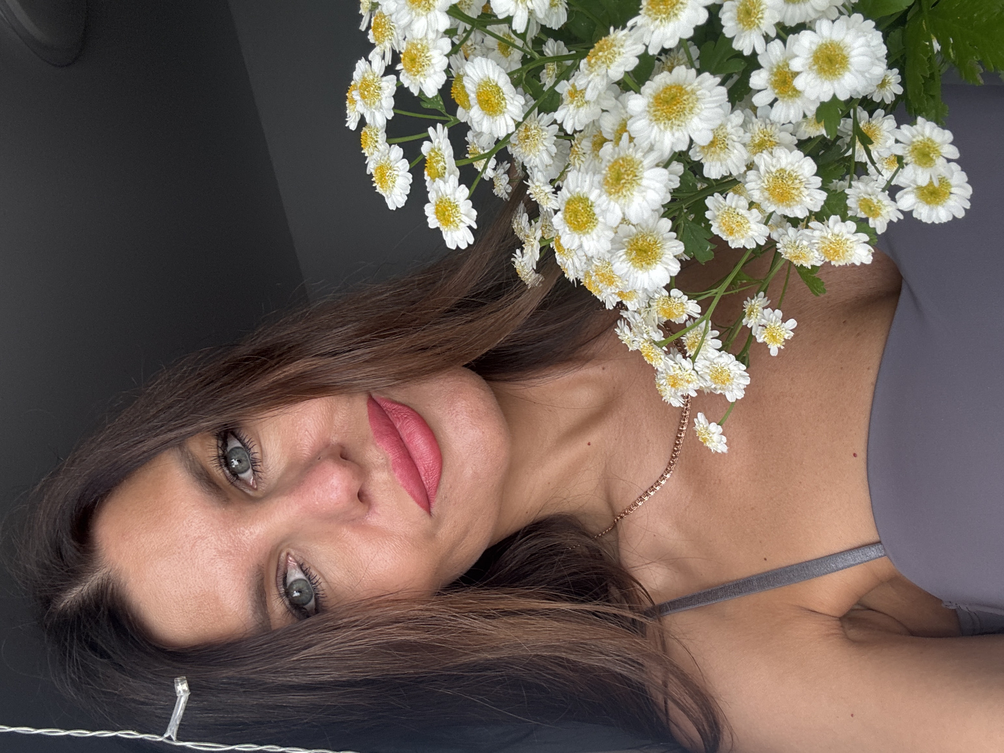 A woman with long brown hair and light-colored eyes looks directly at the camera, her face tilted to the side. She is wearing pink lipstick and has on a grey top. She is holding a bouquet of white daisies with yellow centers. The background is dark and out of focus, with a hint of a string of lights at the bottom of the frame. The overall lighting suggests it could be daytime, possibly indoors given the dark background. The image appears to be a close-up selfie.