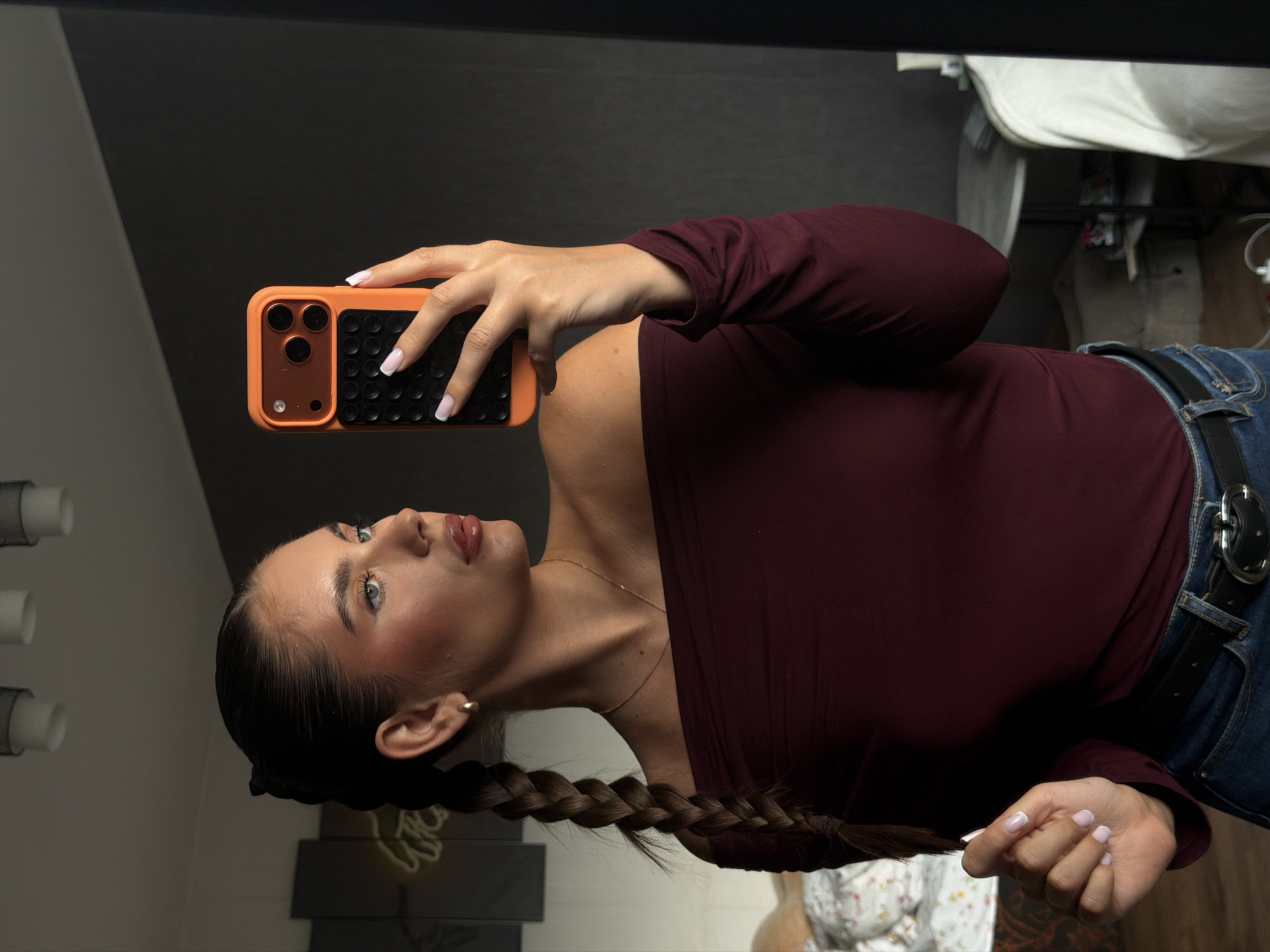 A young woman with light skin, brown hair styled in a braid, and wearing a maroon off-the-shoulder long-sleeved shirt and blue jeans with a black belt, is holding an orange and black phone in her right hand, taking a selfie in a room with a dark gray wall, light fixtures, and a mirror. The location is potentially in Krasnoyarsk, Russia.