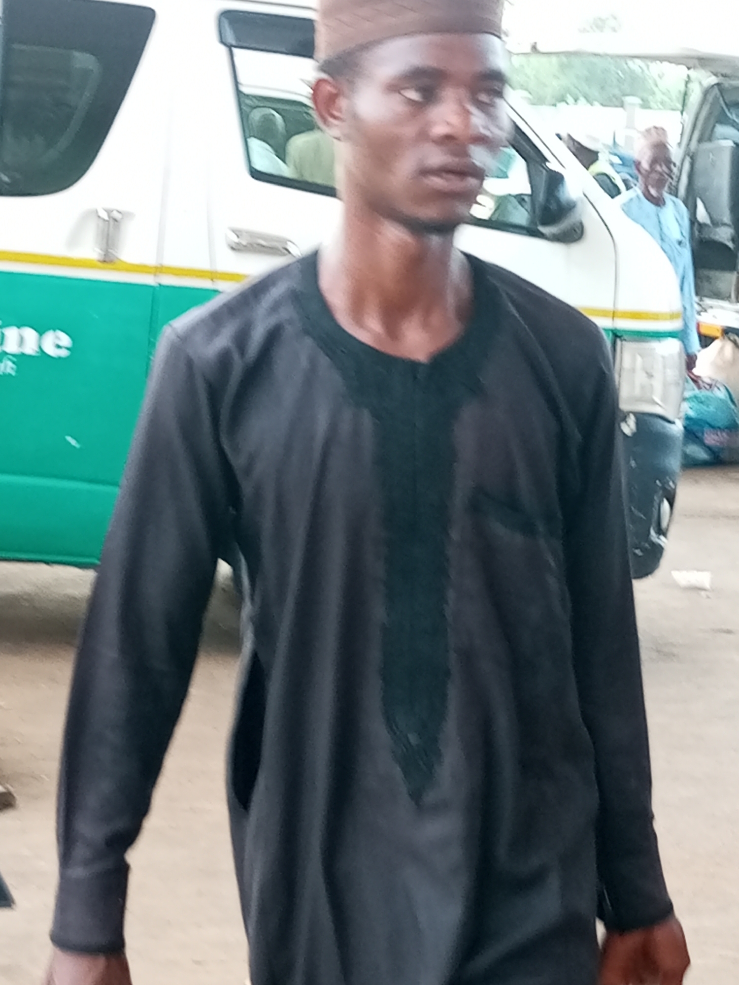 A man in a dark robe and brown cap stands in front of a white and green vehicle, possibly a bus, in Gombe, Nigeria. The vehicle has the word "line" written in white on its side. Another man in a light blue robe is visible inside the vehicle. The ground is a sandy, dirt surface, and in the background, there are bags and other objects, suggesting a market or transport hub. The lighting is natural daylight, and the image is taken at eye level.