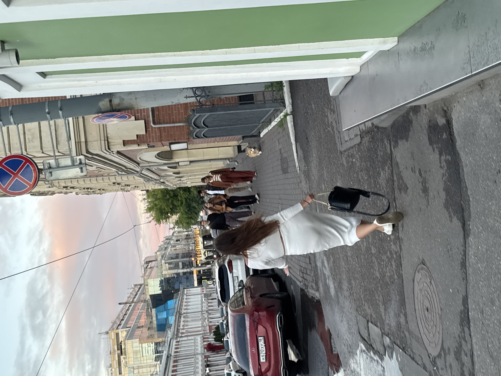 The image depicts a street scene in Nizhny Novgorod, Russia. The foreground shows a woman in a white dress walking away from the camera, carrying a black handbag. She is wearing white sneakers. She is walking on a wet sidewalk next to a parked red car. In the background, several other cars are parked along the street. A group of people in casual clothing are walking on the sidewalk in the distance. The architecture suggests an urban environment with a mix of older and newer buildings. There are also traffic signs visible, including a "No Stopping" sign. The sky is overcast with a hint of pink and orange, suggesting it might be dusk or dawn. The overall atmosphere is calm and ordinary, depicting a typical day in the city. No specific event is evident, and the emotions of the individuals are not discernible. The license plate of the red car is visible and reads "H470XY 18".