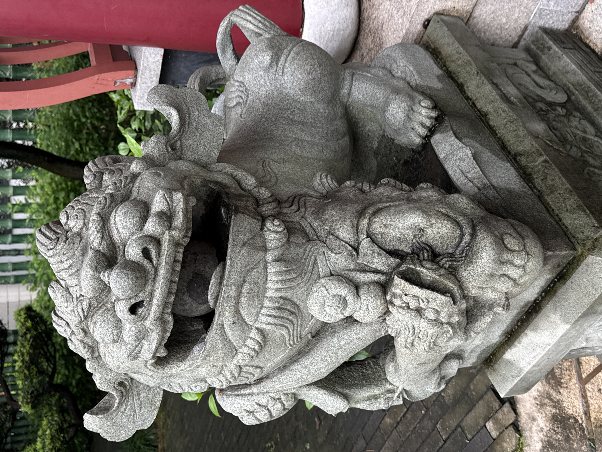 A weathered, grey stone statue of a Foo dog, or lion dog, is positioned outdoors. The statue is intricately carved with a fierce expression, its mouth open to reveal a ball inside. It is seated on a multi-tiered stone pedestal, which is itself situated on a paved area with some greenery visible in the background. To the right, a portion of a bright red cylindrical pillar and a red wooden architectural element are visible. The Foo dog statue appears to be an outdoor guardian figure, likely part of a temple or traditional building in Futian District, China.