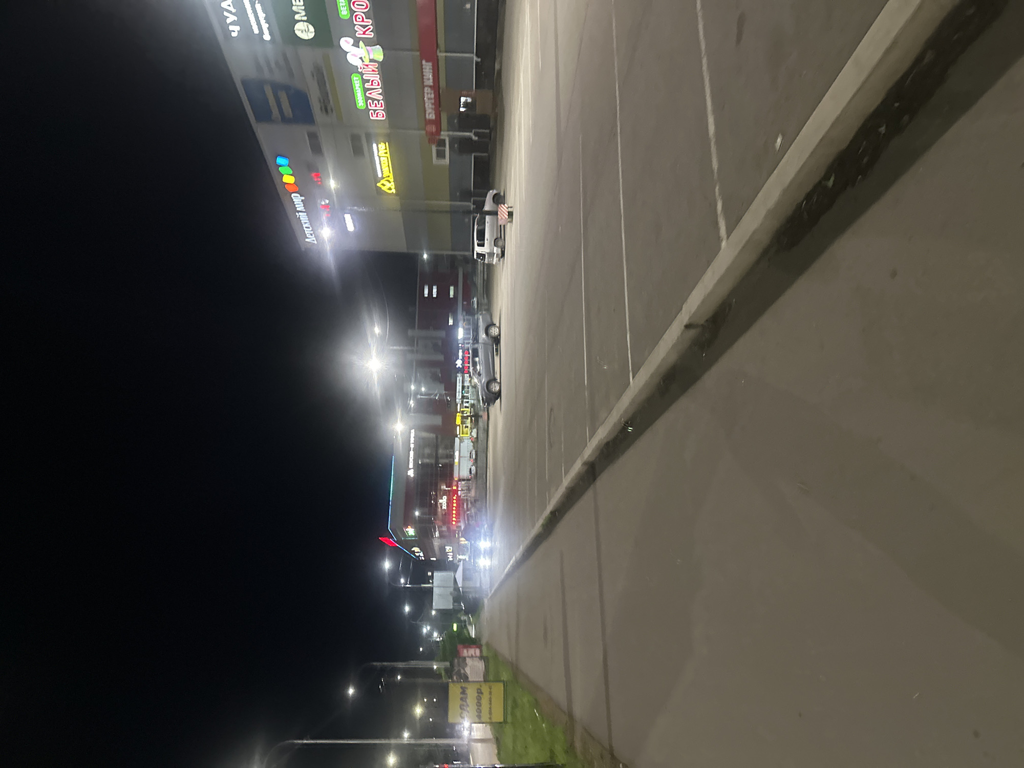 This image depicts a nighttime scene in Birobidzhan, Russia, likely in front of a commercial building or shopping center. The perspective is from street level, looking towards the building.
The primary subjects are the illuminated signs of various shops. One prominent sign displays "ДЕТСКИЙ МИР" (Children's World) with colorful lights. Another sign, also highly visible, reads "БЕЛЫЙ КРОЛИК" (White Rabbit) in large, stylized lettering. There are other signs with text that is partially obscured or difficult to read due to the angle and lighting, but some appear to be in Cyrillic script.
The scene is set in a parking lot or an area adjacent to a roadway, indicated by asphalt paving and painted white lines marking parking spaces. Several vehicles are present, including what appears to be a white minivan or SUV parked in the lot and other cars further down the street, some with their headlights on, suggesting they are either moving or parked. A concrete curb and sidewalk are visible in the foreground, separating the paved area from a grassy verge.
The time of day is clearly night, given the darkness and the artificial illumination from the signs and streetlights. The weather appears clear, with no visible rain or fog. The overall mood is typical of a commercial area at night, with an emphasis on commercial advertising and the availability of services.
Notable details include the brightness of the signs, which stand out against the dark background. The Russian location context is confirmed by the Cyrillic text on the signs. There are no visible people in the image. The activity taking place is likely simply the illumination of the commercial establishments for the night, with vehicles passing through or parked.