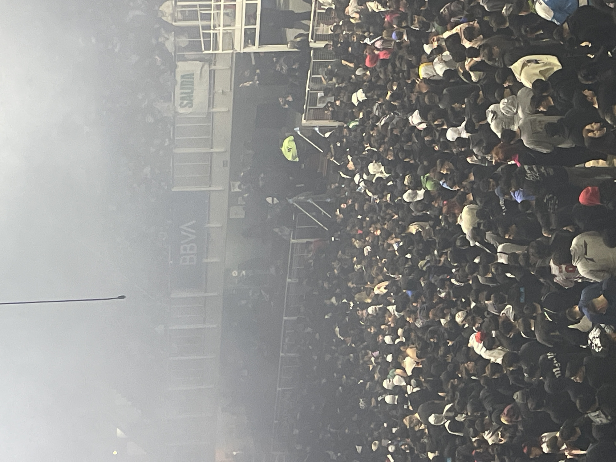 The image shows a dense crowd of people in a stadium, likely in Buenos Aires, Argentina, given the "BBVA" and "SALIDA" signage, typical of Argentine stadiums sponsored by the bank. The crowd is packed together, mostly wearing dark clothing with a few lighter colors mixed in. There's a person wearing a bright yellow jacket standing on the steps, potentially security. The stadium structure is visible on the left, obscured by smoke or fog. The photograph is taken from an elevated position, looking down at the crowd.