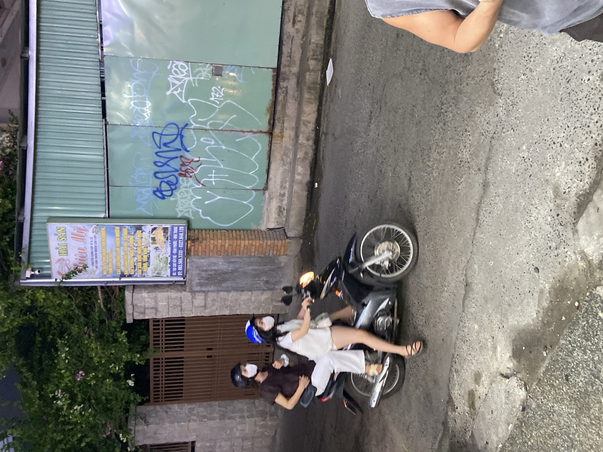 Two individuals, both wearing face masks and helmets, are riding a black motorcycle on a street in Bac Nha Trang Ward, Vietnam. The person in front is driving, wearing a white t-shirt and shorts, and a blue and white helmet. The person in the back is wearing a dark brown top and white pants, with a black helmet. They are both facing forward. The street appears to be paved with asphalt and has some patches of wetness and debris. Behind them, there is a metal gate and a brick wall. A sign with Vietnamese text and images of seafood is mounted on the wall. Further in the background, there is a corrugated metal fence covered in graffiti. The lighting suggests it is either dusk or a heavily shaded area.