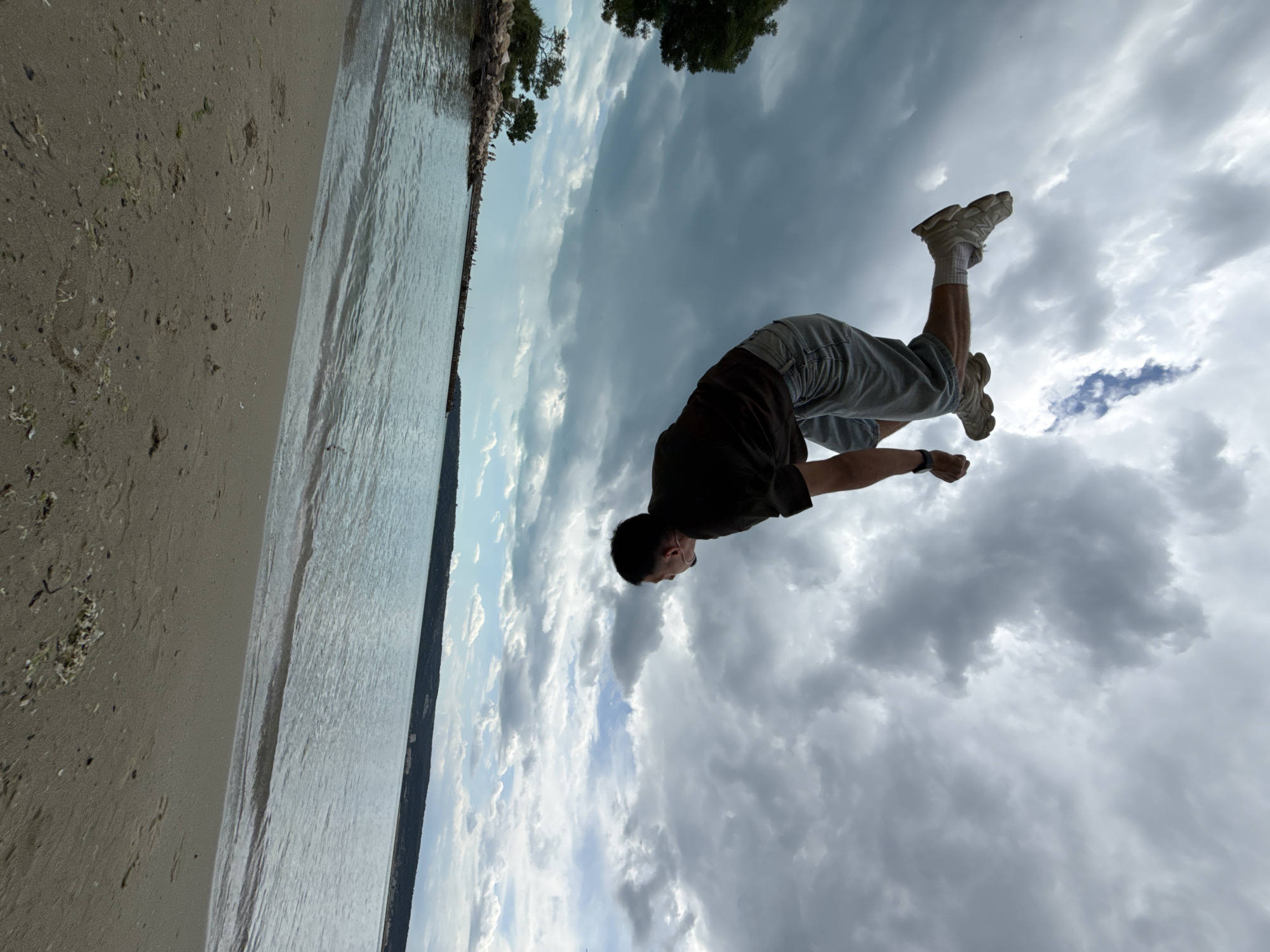 A man is performing a backflip near a body of water. The location is likely a beach in Varna, Bulgaria, considering the sand and water. The sky is overcast. The man is wearing a dark-colored shirt, light-colored shorts, white socks, and white sneakers.