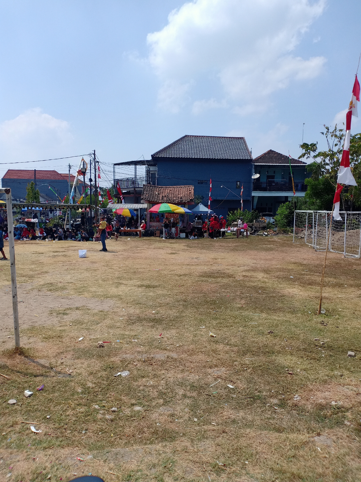 The image depicts an outdoor event, possibly a community gathering or a sports event, taking place in a grassy field in Sumber Tlaseh, Indonesia. The field is dry and partially covered with sparse grass, with visible litter scattered on the ground. In the background, several buildings are visible, including houses with red and blue exteriors, and a larger, dark blue multi-story building. A canopy tent with colorful umbrellas is set up, providing shade to a gathering of people. Several individuals, including adults and children, are present, some seated and others standing. Indonesian flags, red and white, are displayed on poles and strung between structures, suggesting a national celebration or event. Two white soccer goals with nets are set up on the right side of the field. A single pole with a flag is positioned in the foreground on the right. The sky is clear and blue with scattered white clouds.