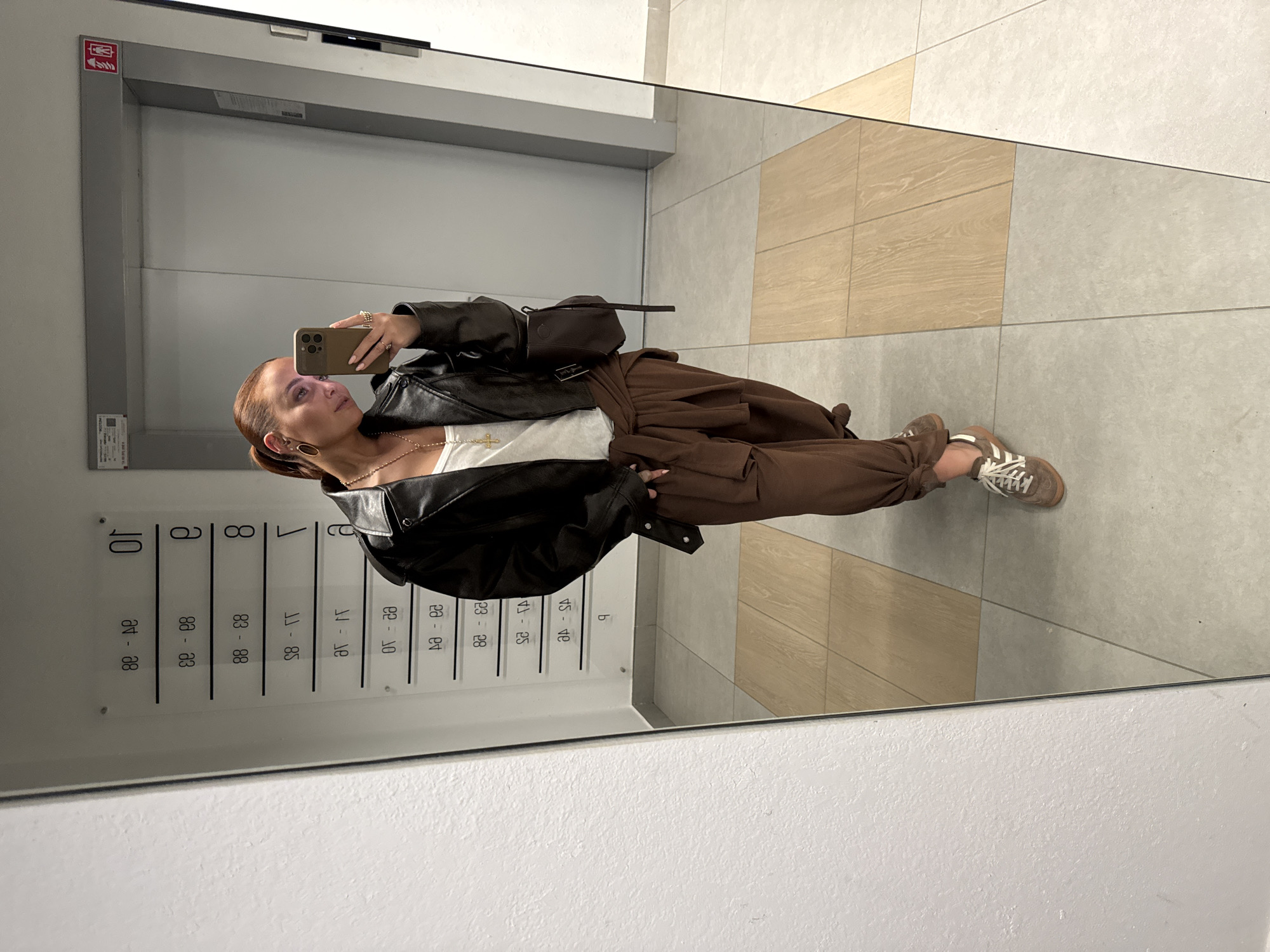 A woman in a black leather jacket and brown pants is taking a selfie in a mirror. The mirror is reflecting a hallway with numbered apartments on the wall. The floor is tiled in neutral tones. The location is likely a building in the city of Izhevsk, Russia, based on the apartment number information.