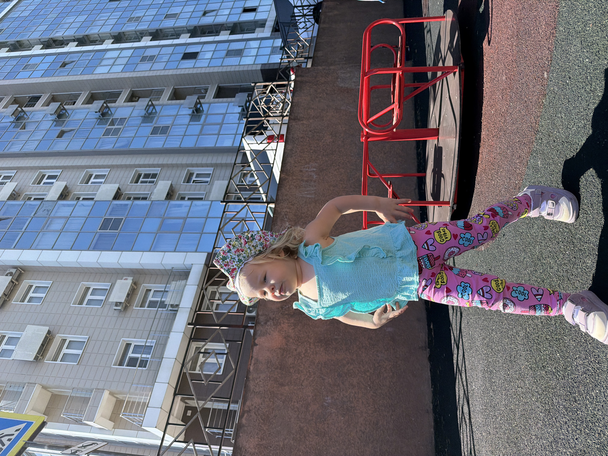 A young girl stands on a playground surface in an urban setting, likely in Sochi, Russia. She has blonde hair and is wearing a colorful floral sun hat, a light blue ruffled top, bright pink leggings with various cartoon designs (including hearts, donuts, cupcakes, and text like "Good Vibes" and "COOL"), and light-colored sneakers. Her expression is neutral as she looks slightly to her left.
The ground she stands on is a dark-grey, textured playground material, and behind her is a reddish-brown wall or building section. To her right, red metal playground equipment, possibly a small slide or climbing frame, is visible. In the background, a tall, modern building with many reflective blue-tinted windows rises upwards. Several windows are open, and multiple air conditioning units are mounted on its exterior walls. A dark metal fence or railing separates the playground from the building structure.
The scene is well-lit by bright sunshine, casting distinct shadows that suggest it is mid-day to early afternoon on a clear, sunny day. A partially obscured blue and white road sign is visible in the bottom-left corner of the frame.