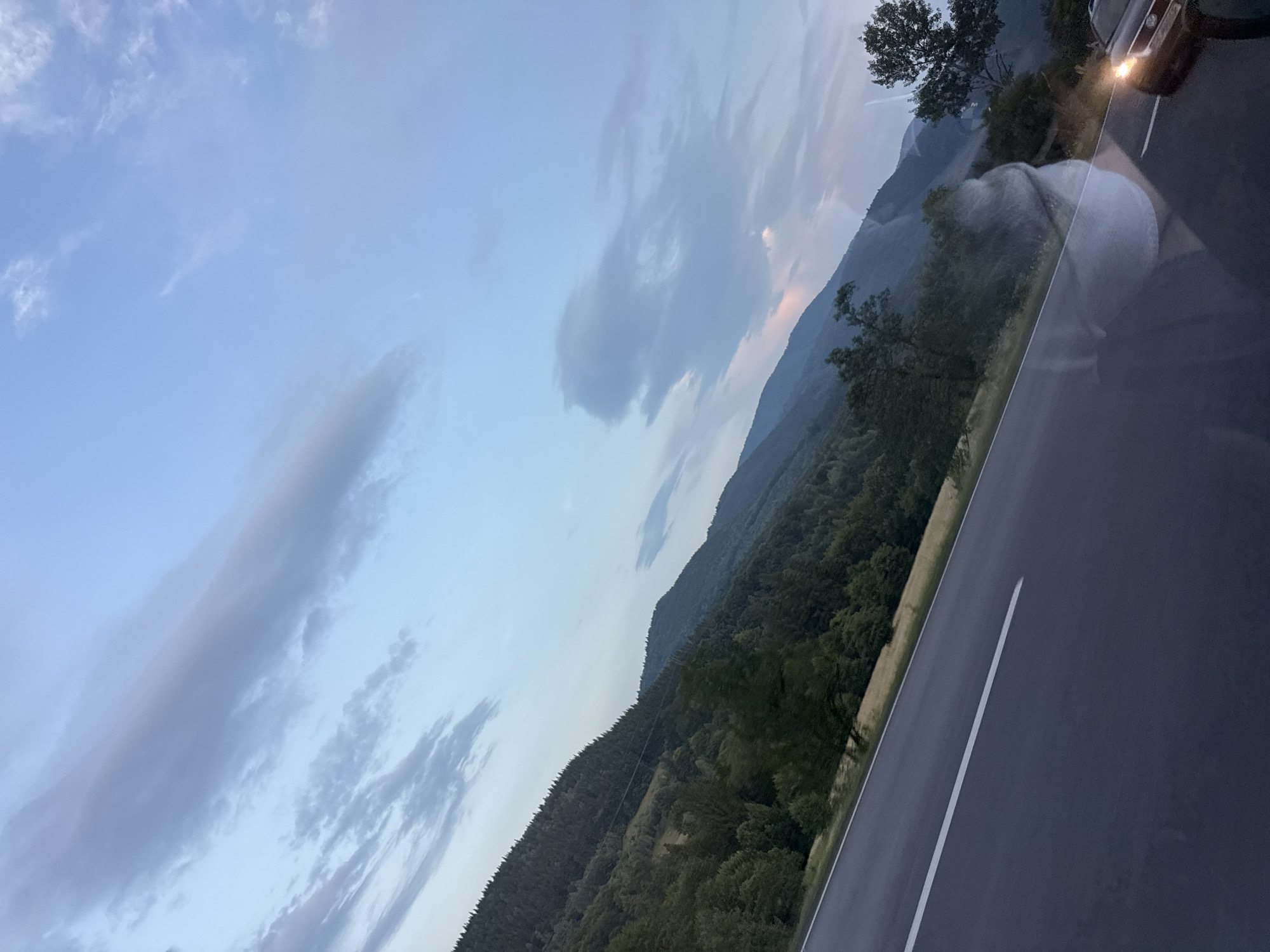 This is a tilted, eye-level, outdoor, daytime, full shot of a winding road in Brețcu, Romania. The sky is a soft blue with scattered clouds. Mountains covered in dense green trees rise from the right side of the frame, sloping down towards the left. A paved road with a white stripe runs diagonally from the bottom left to the upper right of the image. A car with its headlights on is visible on the right side of the road, approaching the viewer. The reflection of a person is visible on the car's windshield.