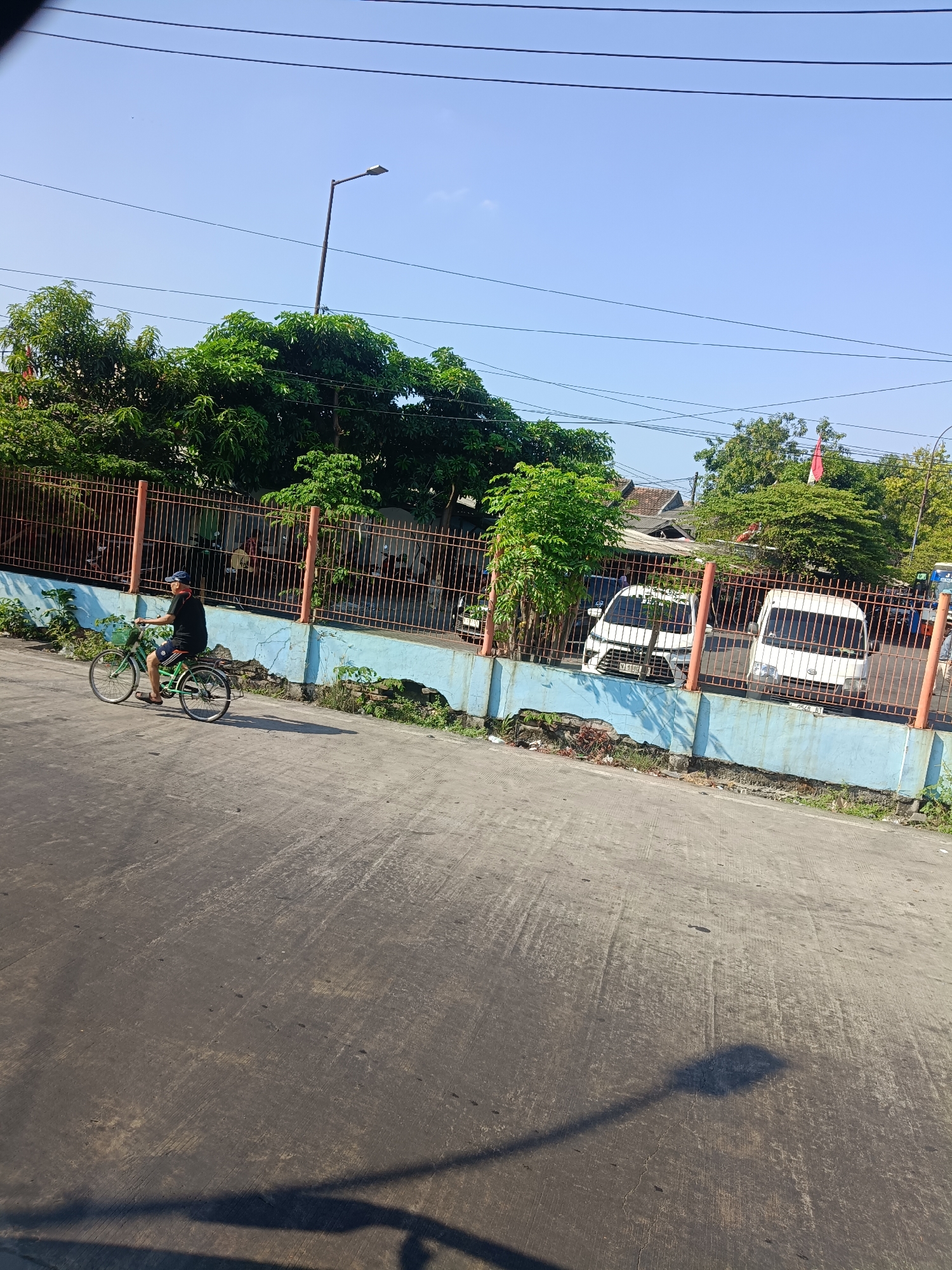 A man is riding a bicycle on a paved road in front of a fenced area. The man is wearing a black t-shirt and shorts, and a blue cap. The bicycle is green and has three wheels. Behind the fence, there are trees, two white vans, and a street lamp. The sky is clear and blue. The scene is likely located in the city of Kedungrejo, Indonesia.