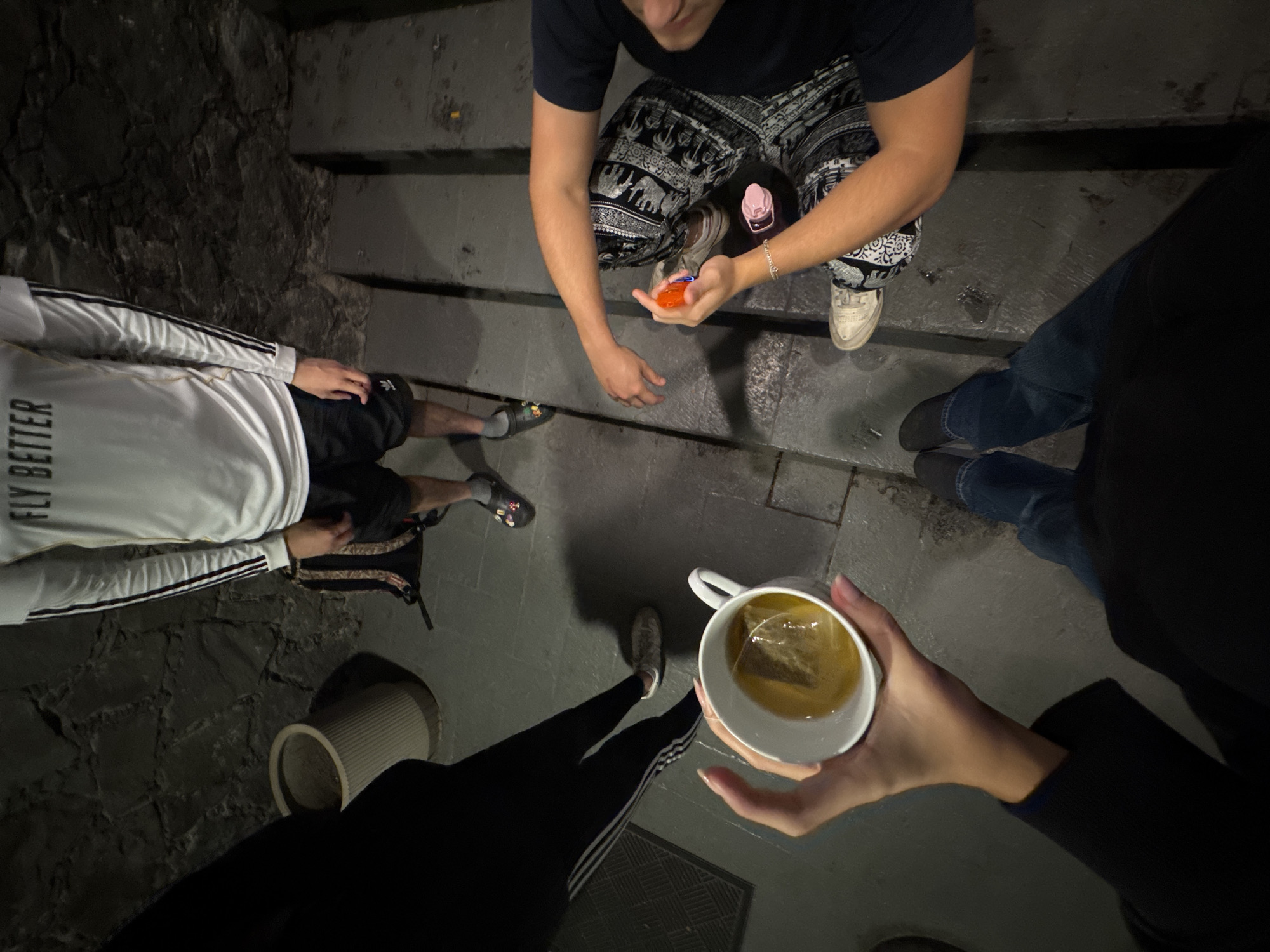An overhead, low-light photograph captures a casual nighttime gathering of at least four individuals on a stone and concrete surface, possibly a set of steps or a pathway in Mexico City. The scene suggests an outdoor or dimly lit semi-enclosed space, with an uneven, dark stone wall visible on the left.
In the center, a person crouches, wearing a dark t-shirt, black and white patterned pants (featuring elephant motifs), and white sneakers. They hold a small, translucent orange object, possibly a toy or container, and a pink water bottle rests beside them. To the left, another person stands wearing a white long-sleeve shirt with the text "FLY BETTER" on the back, black shorts, and flip-flops. A backpack is visible near their legs. On the far right, only the lower legs of a person in blue jeans and dark shoes are visible.
In the immediate foreground, a person holds a white mug filled with tea, identifiable by a tea bag steeping within. This individual wears black leggings with white stripes and white sneakers. Their hands, with neatly manicured nails, cradle the mug.
The soft, ambient light suggests an evening or nighttime setting. The group appears to be engaged in a relaxed social interaction. A white, ribbed planter or pot is partially visible in the bottom-left corner of the frame.