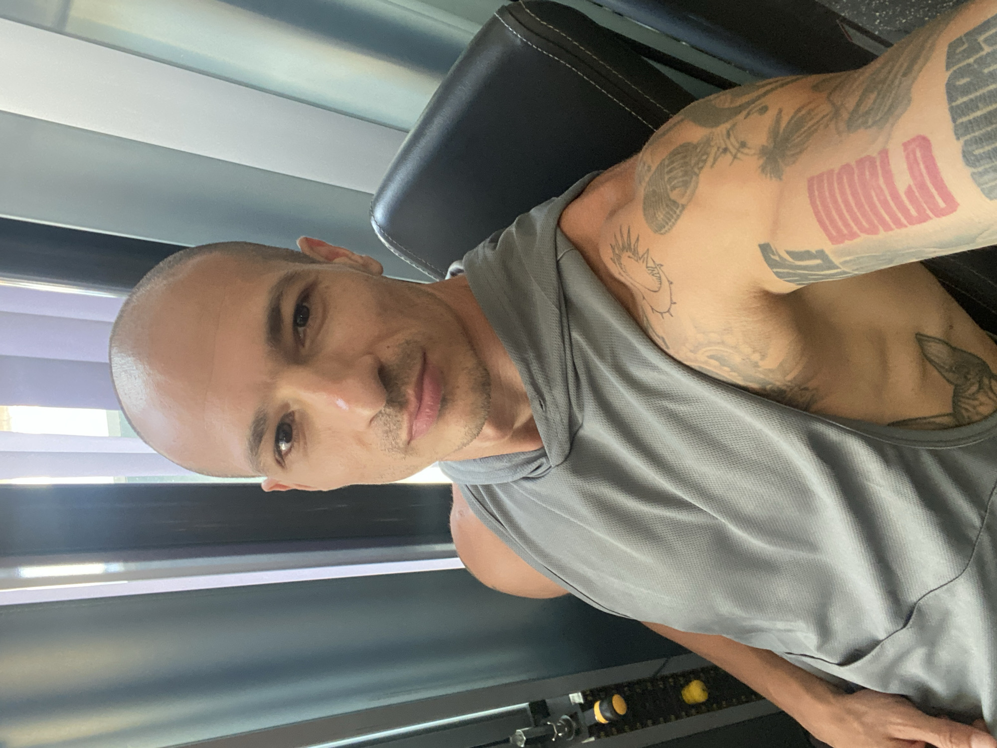 In a gymnasium, a bald man with tattoos on his arms and a gray tank top is taking a selfie. He is seated on a black gym seat, with a gym machine to his left and a window with vertical blinds to his right. The image provides no information about the city of Pleven, Bulgaria.