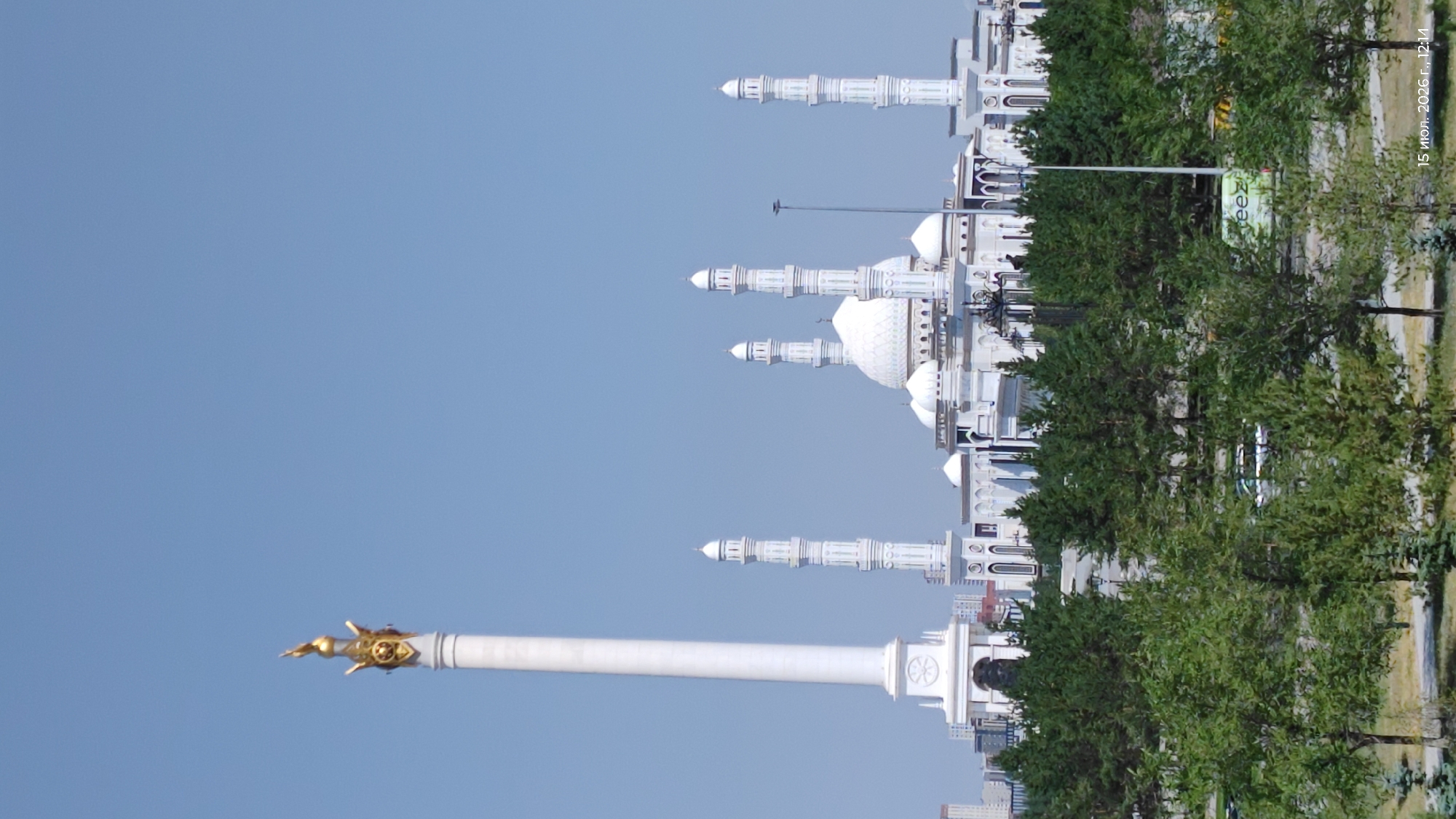 A wide-angle, eye-level exterior shot captures a clear, bright day in Astana, Kazakhstan, with a prominent monument and a mosque as the main subjects. On the left, a tall, white, fluted column monument topped with a golden sculpture rises majestically against a pale blue sky. To its right, a large, white mosque complex dominates the mid-ground. The mosque features multiple domes, with a central, larger dome surrounded by smaller ones. Several tall, slender minarets, also white, punctuate the mosque's structure, with four visible in a row. In the foreground, a dense line of various green trees, primarily pine and deciduous varieties, creates a natural barrier, partially obscuring the lower portion of the buildings and a street with a white vehicle and a green truck visible behind the trees. The date and time stamp "15 июл. 2026 г., 12:14" is present in the lower right corner of the image.