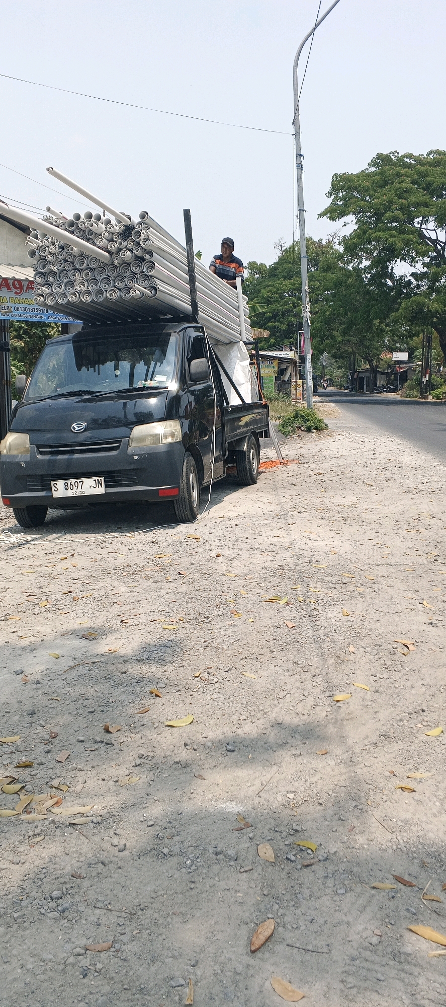 In Karangbinangun, Indonesia, a black Daihatsu Gran Max pickup truck is parked on a gravelly dirt road beside a paved road. The truck bed is filled with a large load of silver-colored pipes stacked horizontally, some appearing to be PVC. A man wearing a horizontally striped t-shirt and a dark baseball cap stands on top of the pipes, looking towards the right. A white tarp is also visible in the truck bed. The scene is outdoors on a bright day with a clear sky. Trees line the paved road in the background, and a street light is visible on the right side of the image. A sign with Indonesian text is partially visible on a building to the left of the truck. The ground is covered with gravel and scattered dry leaves. The license plate of the truck reads "S 8697 JN".
