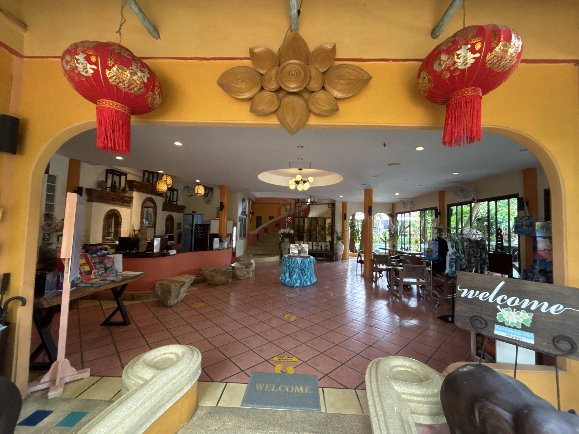 This image depicts the entrance and lobby of a building, likely a guesthouse or small hotel, in Chalong, Thailand. The architecture features warm yellow walls and arched doorways. Two large red Chinese lanterns with gold embellishments hang prominently from the ceiling near the entrance. In the center of the image, a wooden, lotus-shaped decorative element is mounted on the wall.
The lobby area is spacious with tiled floors. Several pieces of furniture are visible, including tables, chairs, and a reception desk. A blue tablecloth covers a round table in the center of the room. Various decorative items are scattered throughout, such as stone sculptures and potted plants. A "Welcome" mat is placed near the entrance, and social distancing markers are visible on the floor. In the background, a staircase leads to an upper level, and large windows offer a glimpse of lush greenery outside. A rack of clothing and souvenirs is displayed to the right, and a wooden sign with the text "welcome, Aohelong villa, Phuket" is also present. No people are actively visible in the scene. The overall impression is one of a welcoming, tropical establishment.