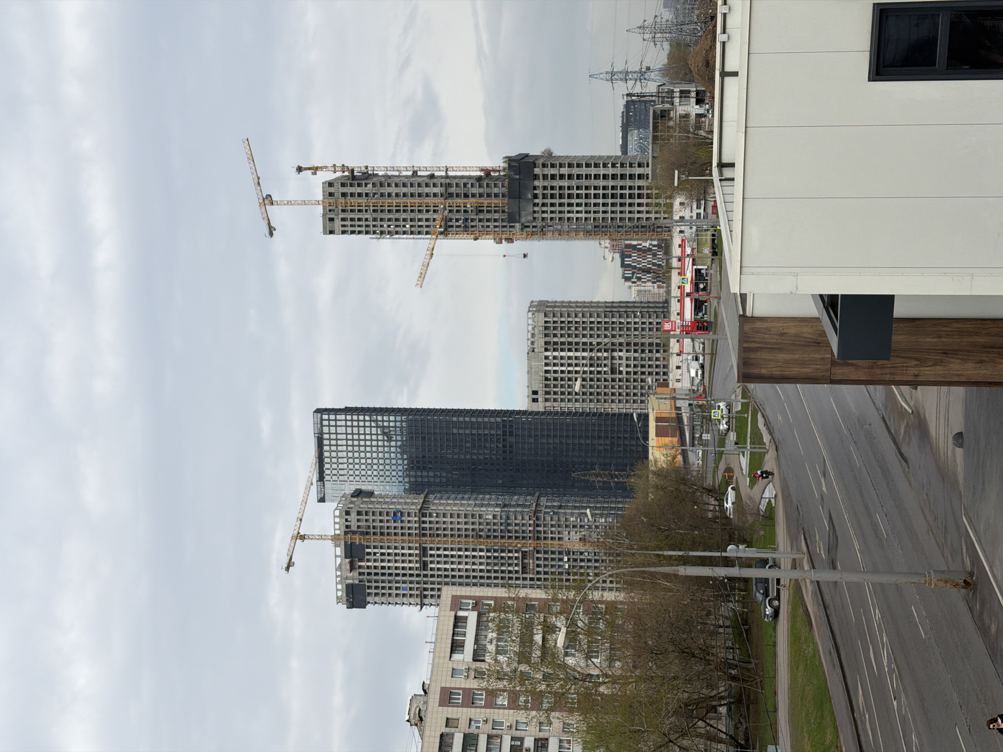 The image depicts a street in Moscow, Russia with several high-rise buildings under construction in the background. The buildings are skeletal structures with multiple cranes attached, indicating ongoing construction. In the foreground, a street stretches away from the viewer, flanked by bare trees and streetlights. The road surface appears damp, possibly from recent rain. There is pedestrian and vehicle traffic. To the right, there is the side of a building, providing context for the perspective of the shot. The sky is overcast.
