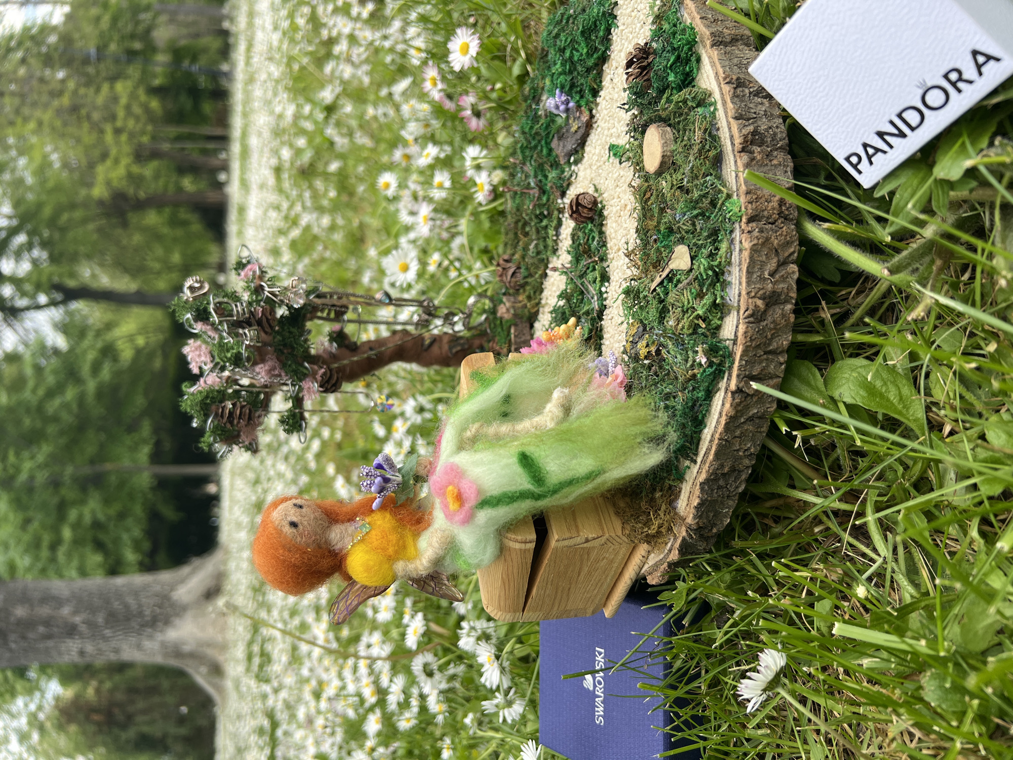 This image, potentially taken in Burgas, Bulgaria, shows a small, crafted fairy scene placed outdoors on a field of daisies and grass. The scene includes a felted figure with orange hair, a yellow body, and green clothing, resting on a miniature wooden bench. Adjacent to the fairy scene is a decorative tree made of twigs and pine cones. The entire setup is placed on a cross-section of a tree trunk that serves as the base. A miniature pathway and moss decorations add to the scene. Two product boxes, one displaying "PANDORA" and the other "SWAROVSKI," are situated near the display, suggesting a connection to these brands. A tree trunk is partially visible in the background. The composition suggests the image was taken outdoors in natural daylight.