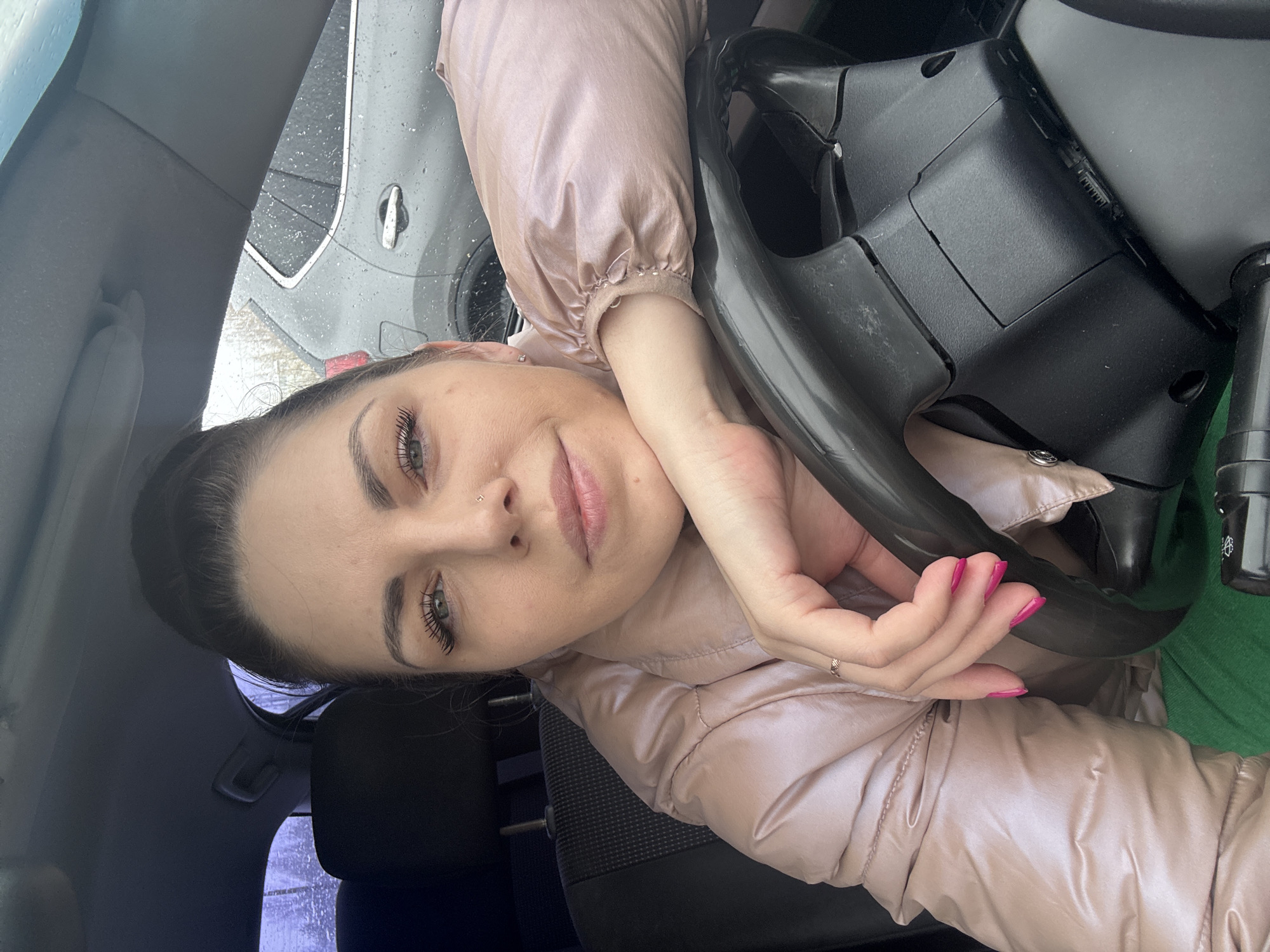 A young woman is captured in a portrait-style shot from inside a car, looking directly at the camera with a calm and slightly smiling expression. Her dark hair is pulled back, revealing light-colored eyes (appearing green or blue-green), and she wears natural-looking makeup, including mascara and a light lip color. She has a small stud piercing on her left nostril and a matching earring on her left earlobe.
She is dressed in a shiny, light pink or rose gold-colored puffy jacket, with a vibrant green garment visible beneath it on her right side. Her right hand, adorned with a small ring on the ring finger and bright pink nail polish, holds the steering wheel. Her left hand/arm is bent, resting on the steering wheel, providing a relaxed pose for her head.
The setting is the interior of a car, with the black steering wheel prominent in the foreground. Parts of the dashboard and a car seat are also visible. Through the car window on the right, the exterior of another vehicle, possibly dark grey, is seen covered in water droplets, indicating rainy or recently rainy weather. The background further suggests an outdoor setting, perhaps a street or parking lot. The lighting appears to be natural daylight, likely overcast. No visible text is clearly legible in the image. The context provided for the location is Shevlyakovo, Russia.