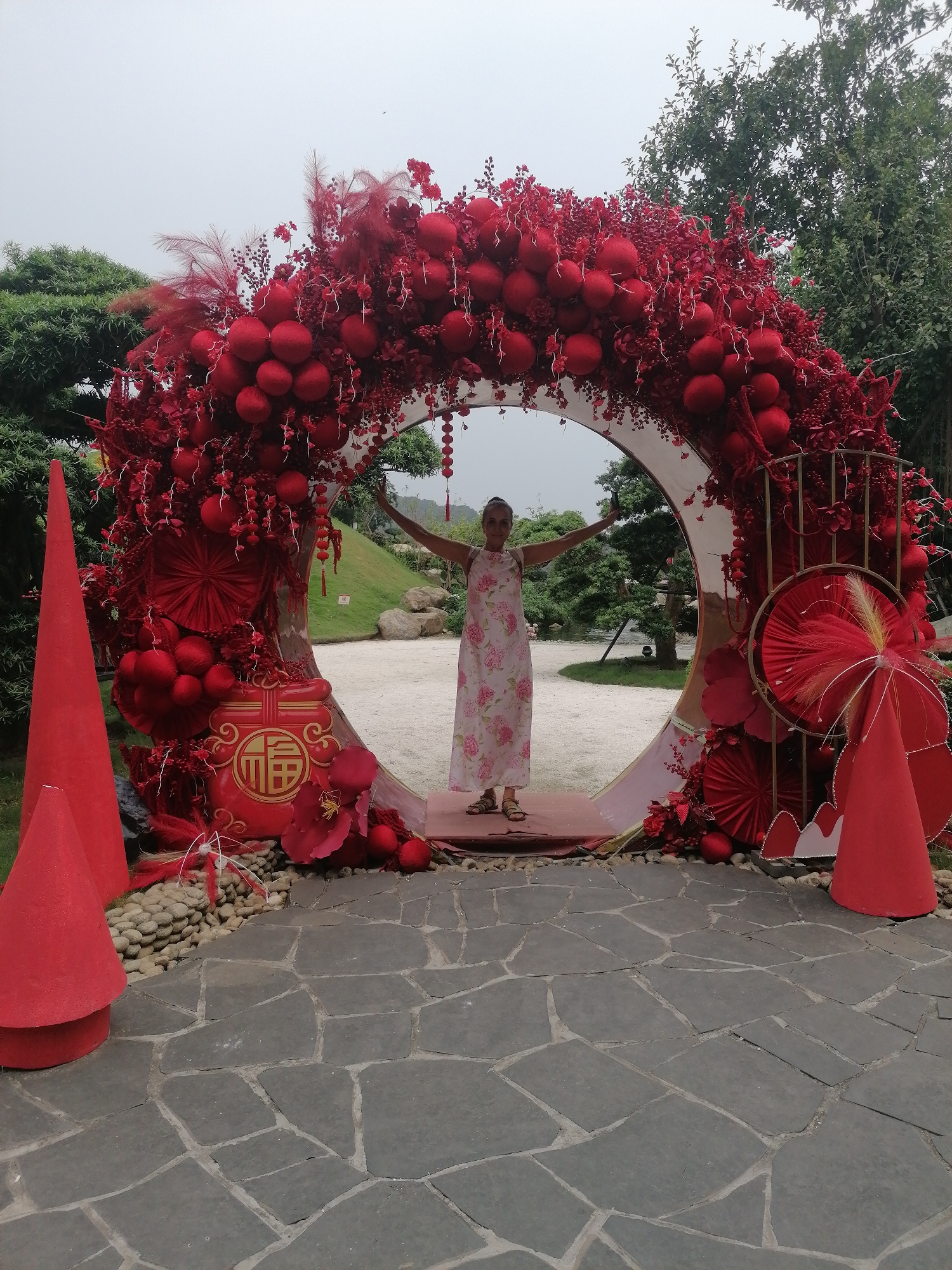 A person stands within a circular, elaborately decorated archway in Hong Gai Ward, Vietnam. The archway is adorned with red spheres and floral arrangements. The person is wearing a light-colored dress with a floral pattern and has their arms outstretched. Two red conical structures flank the archway at the base. The path leading to the archway is paved with irregularly shaped stones. The area surrounding the display includes landscaping with greenery. The sky is overcast.