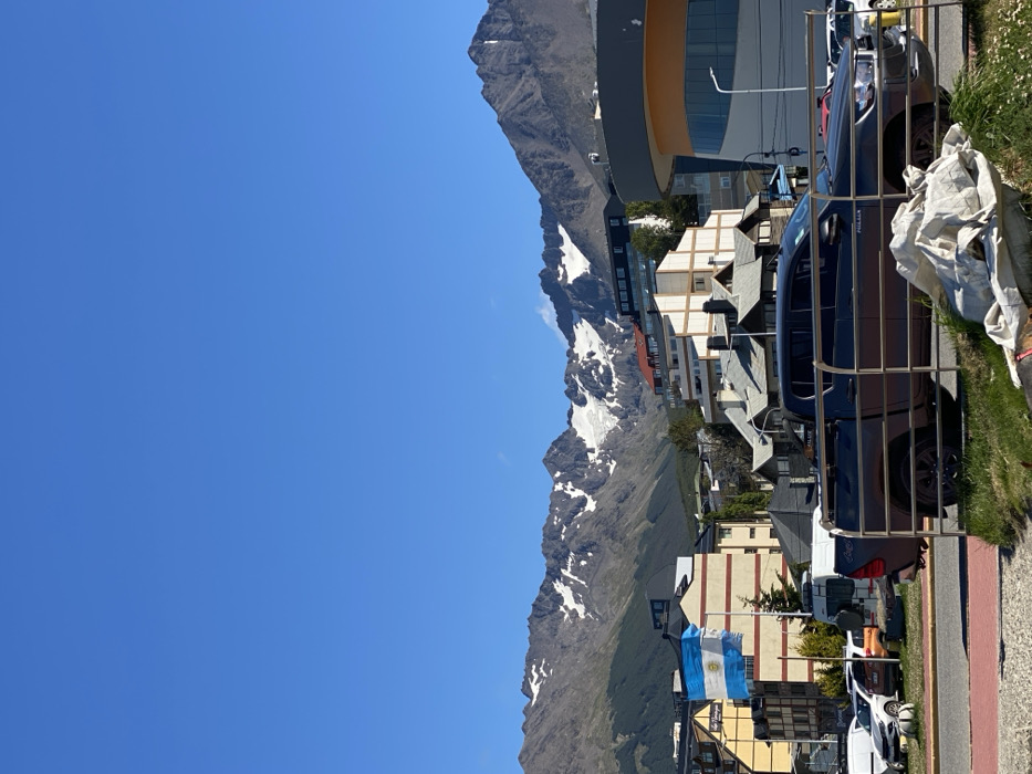 The image depicts a vibrant, sunny day in what appears to be a town nestled at the base of snow-capped mountains. The perspective is from street level, looking towards the mountains that rise prominently in the background.
The sky occupies a significant portion of the left side of the frame, showcasing a clear, brilliant blue with no visible clouds. The sun is high, indicating either late morning or early afternoon, casting bright light over the entire scene.
Dominating the mid-ground are rugged mountains, their peaks and upper slopes still covered with patches of white snow or glaciers, contrasting with the dark, rocky terrain and some sparse green vegetation at lower altitudes. The mountains stretch across the width of the image, forming an imposing natural backdrop to the town.
In the foreground and mid-ground, a collection of buildings forms a town or settlement. The architecture appears modern or relatively recent, with multi-story structures featuring various shades of beige, brown, and grey. Some buildings have flat roofs, while others show sloped, dark-tiled roofs. One notable building on the right side has a curved, modern facade with large windows, and a distinctive orange-brown band running along its upper portion.
A street runs in the immediate foreground, parallel to the viewer's perspective. It has a paved surface, part of which is reddish-brown, possibly a bike lane or decorative strip. To the right of the street, there's a patch of green grass. A dark metal fence, possibly a guardrail, runs along the edge of the street on the right, partially obscuring vehicles parked behind it.
Several vehicles are visible. Behind the fence, a dark-colored pickup truck is parked, with the word "HILUX" visible on its tailgate, indicating it's a Toyota Hilux. Other cars, including white and grey sedans or SUVs, are parked further down the street. A large white tarpaulin or canvas material is bunched up on the grass next to the fence on the far right.
A prominent detail in the lower mid-ground is a light blue and white flag, characteristic of the Argentine flag, flying from a pole attached to one of the buildings. Its colors are vibrant against the blue sky and buildings, suggesting the location is in Argentina.
Overall, the scene conveys a sense of a lively town situated in a breathtaking natural environment, likely a popular spot for tourism given the mountain views and clear weather.