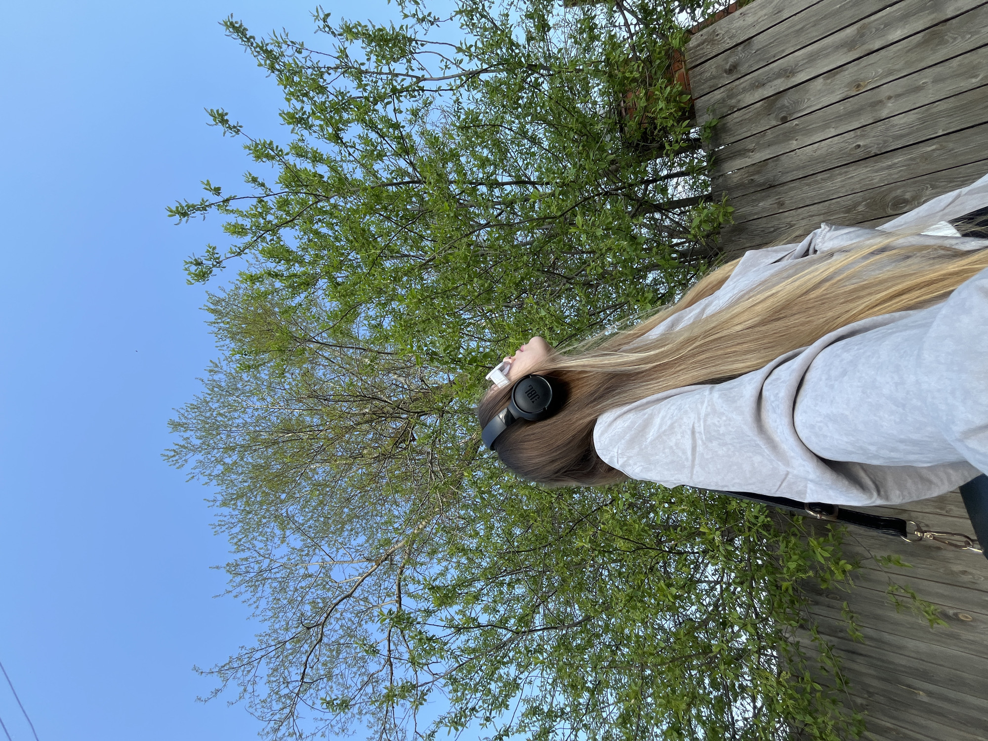 A young woman is looking up into the branches of a tree in Фоки, Russia. The sky is clear and blue, and she is wearing headphones. A wooden fence is visible behind her.