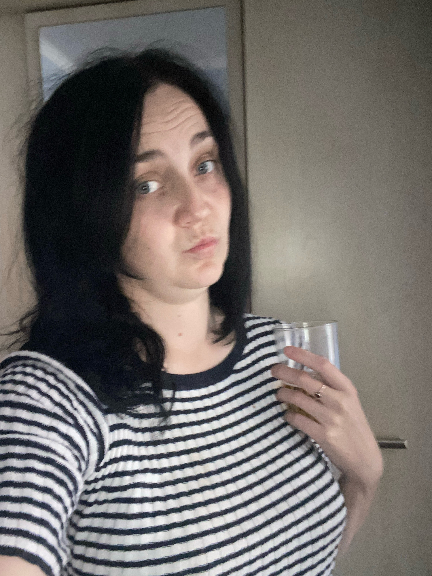 An individual with black hair and blue eyes, wearing a black and white striped shirt, holds a clear glass of amber liquid. The individual's right hand, adorned with a ring, is near the glass. The background appears to be an indoor setting with a light-colored wall and a door handle.